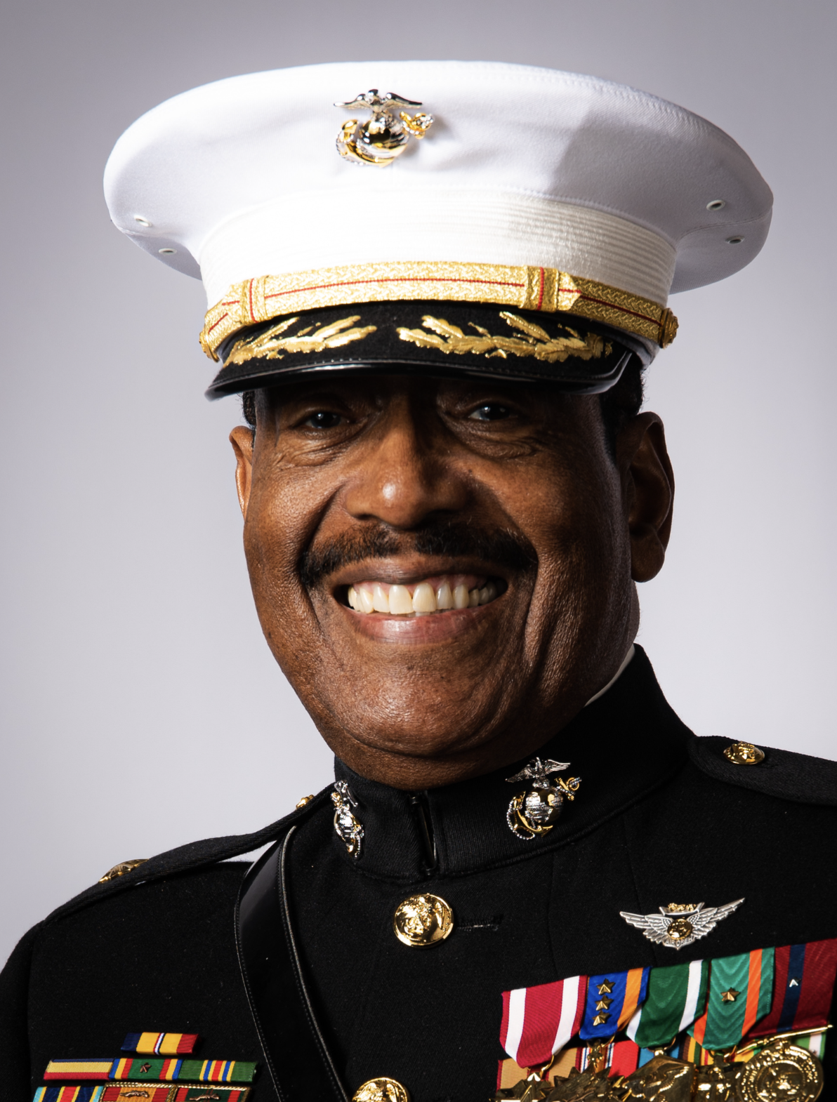 A smiling man in a U.S. Marine dress uniform, wearing a white hat with a gold emblem, black jacket with medals, ribbons, and badges, and a background with neutral gray gradient.