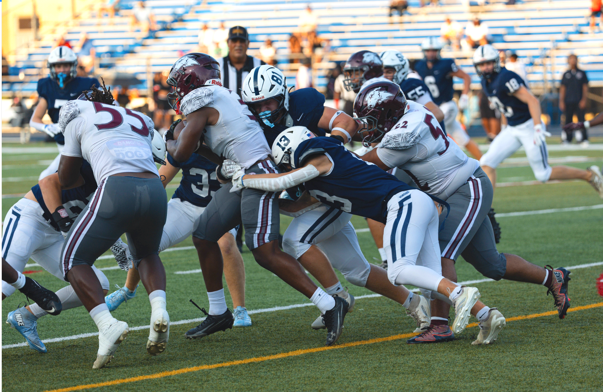 American football players in a tackle during a game on a football field with a referee and spectators in the background.
