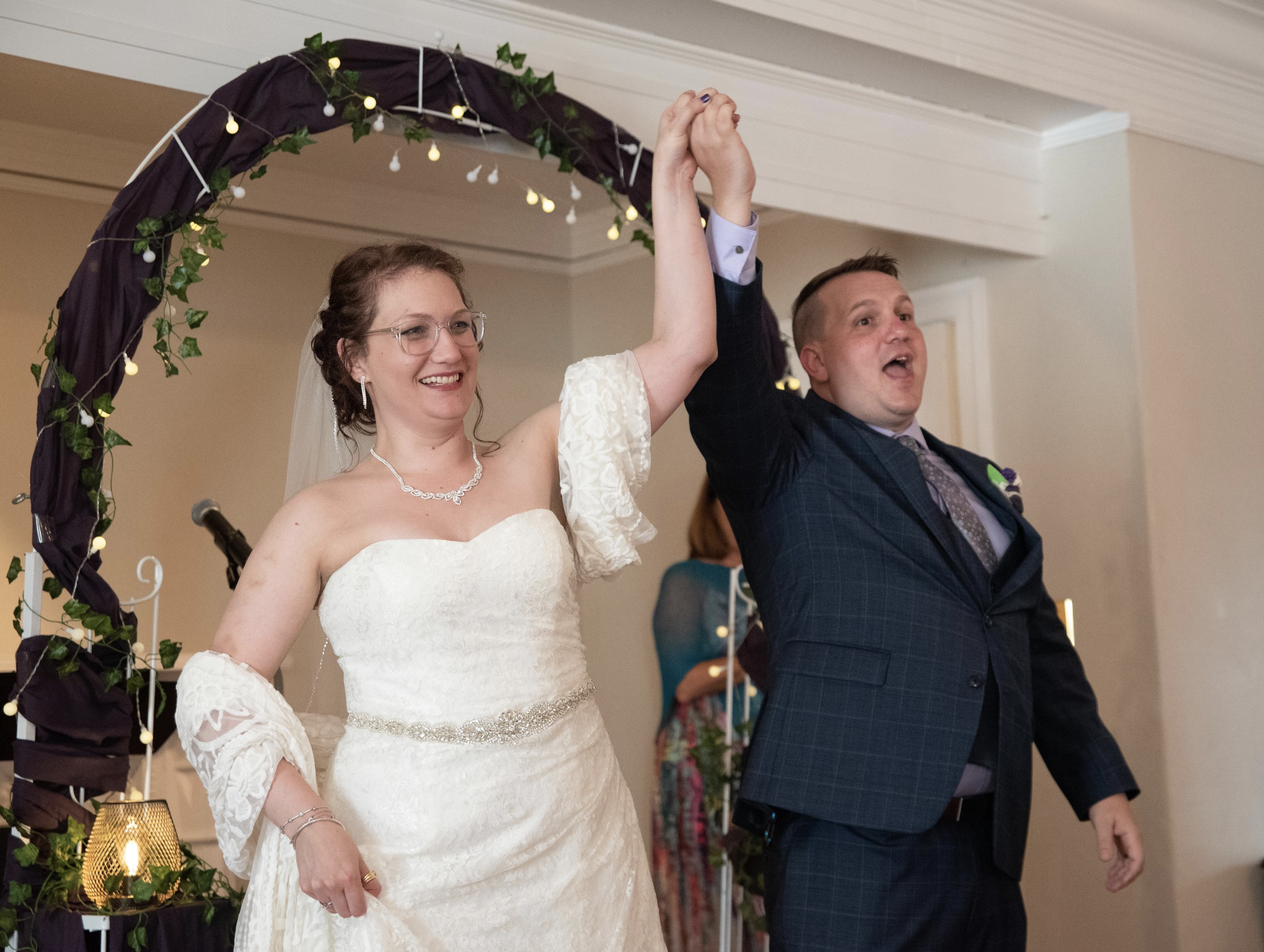 A bride and groom at their wedding reception, holding hands and smiling under a decorated arch with string lights and greenery.