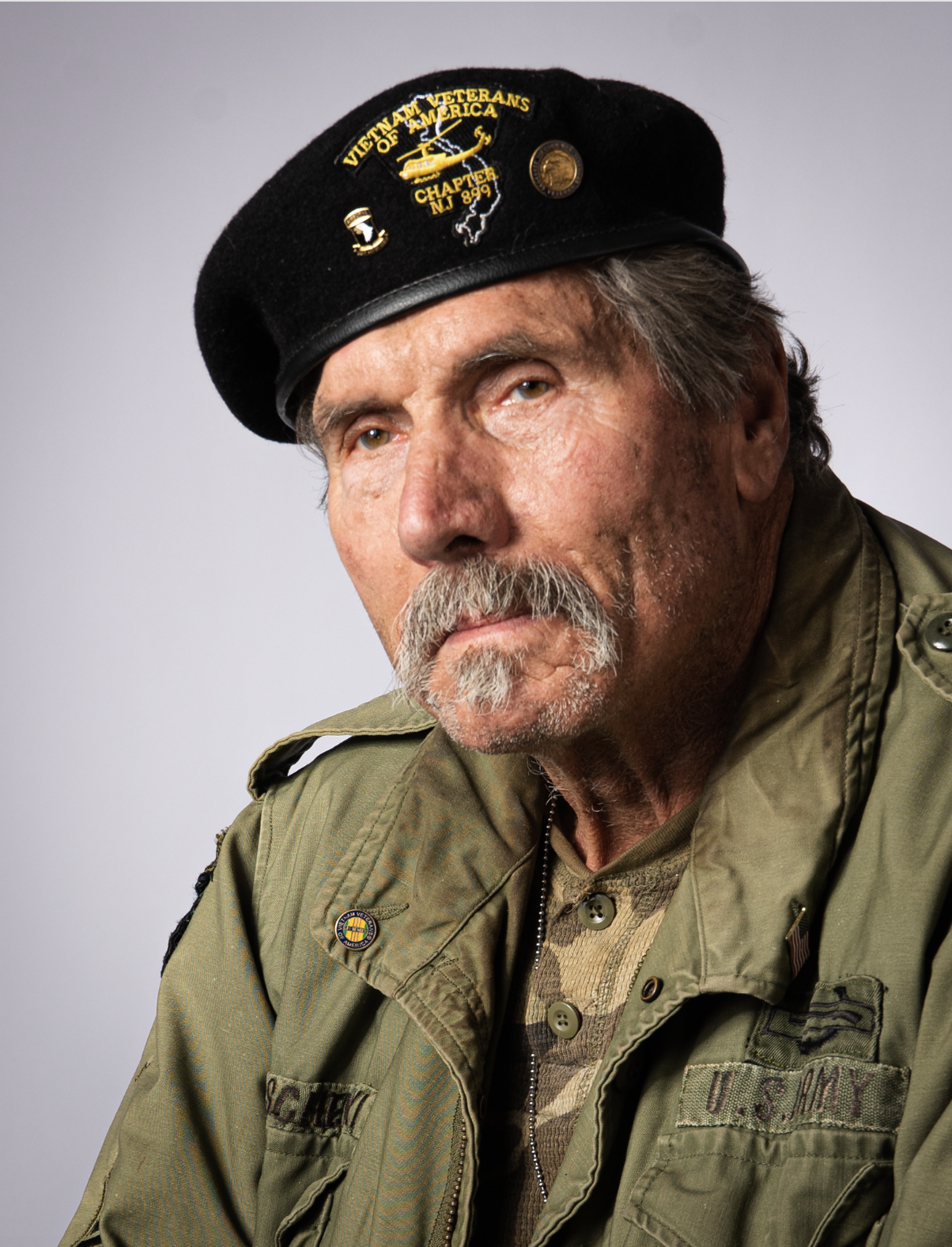Close-up portrait of an elderly man wearing a black Veterans of America beret with patches and pins, dressed in a military green jacket with U.S. Army patches, against a plain gray background.