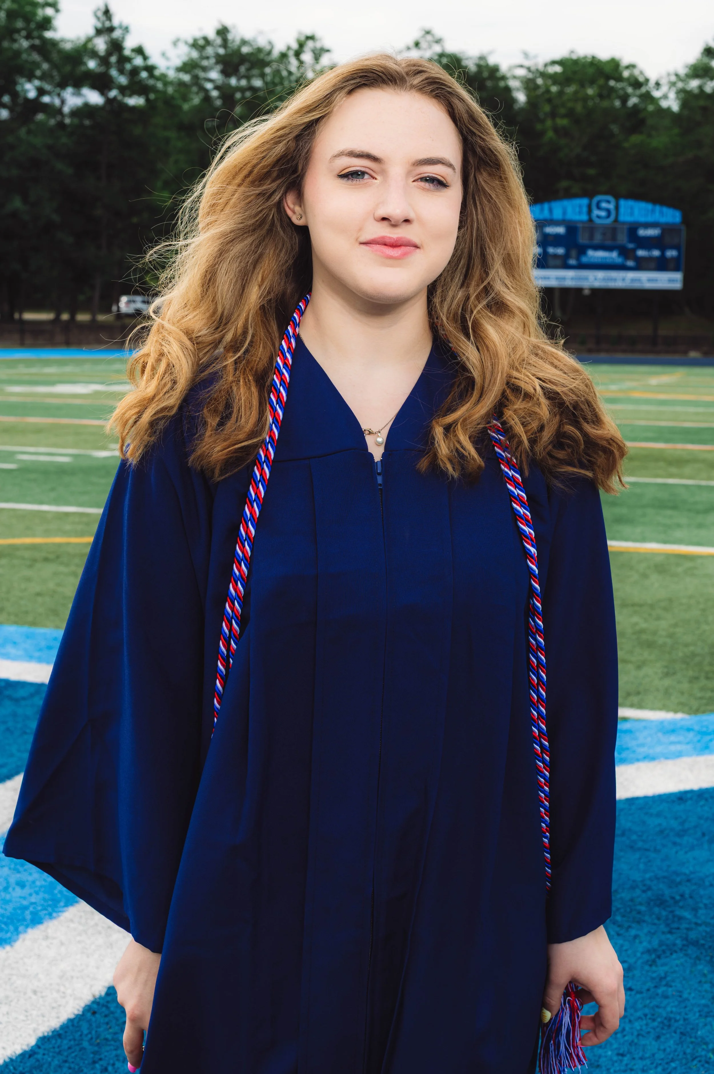 Young woman in a navy graduation gown and honor cords standing on a sports field with a scoreboard and trees in the background.