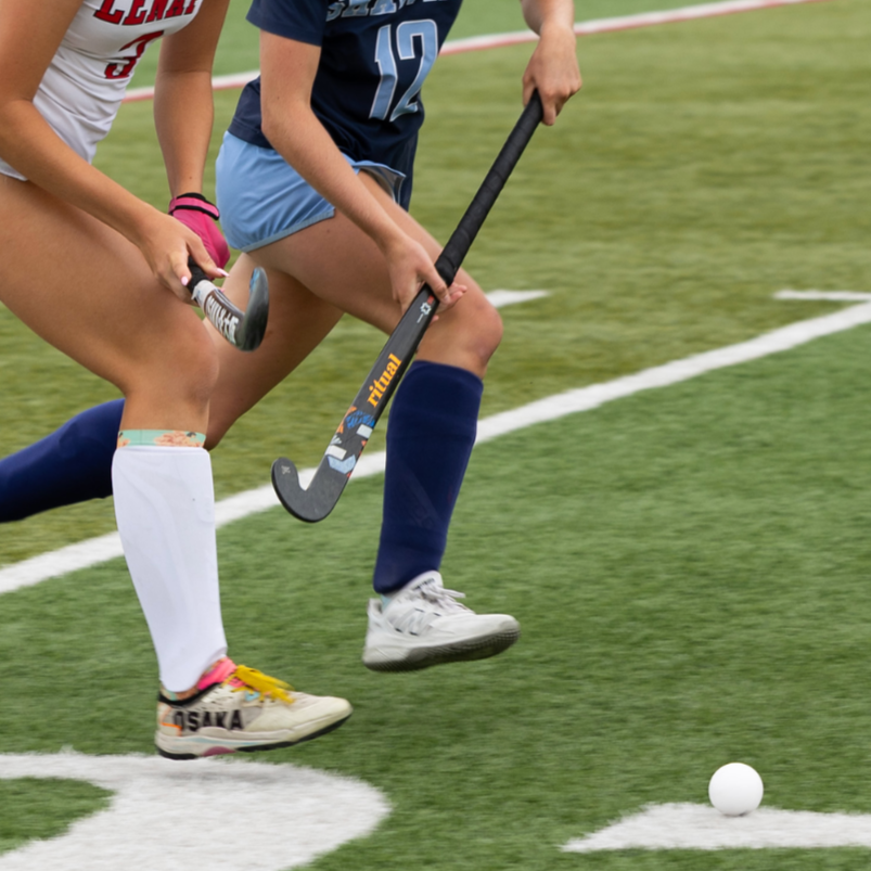 Two female field hockey players battling for control of the ball on a green turf field.