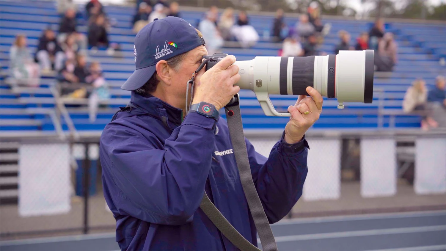 A person wearing a blue jacket and matching baseball cap taking a photograph with a large professional camera featuring a white telephoto lens, at an outdoor event with spectators seated in blue bleachers in the background.