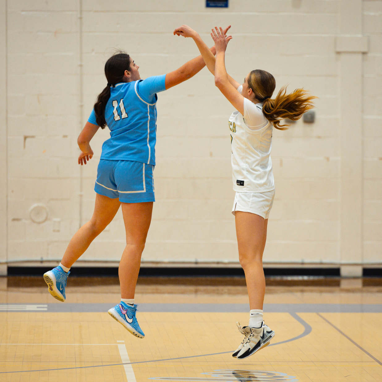 Two female basketball players are jumping and reaching for the ball in an indoor court, one wearing a blue uniform with the number 11, the other in a white uniform.