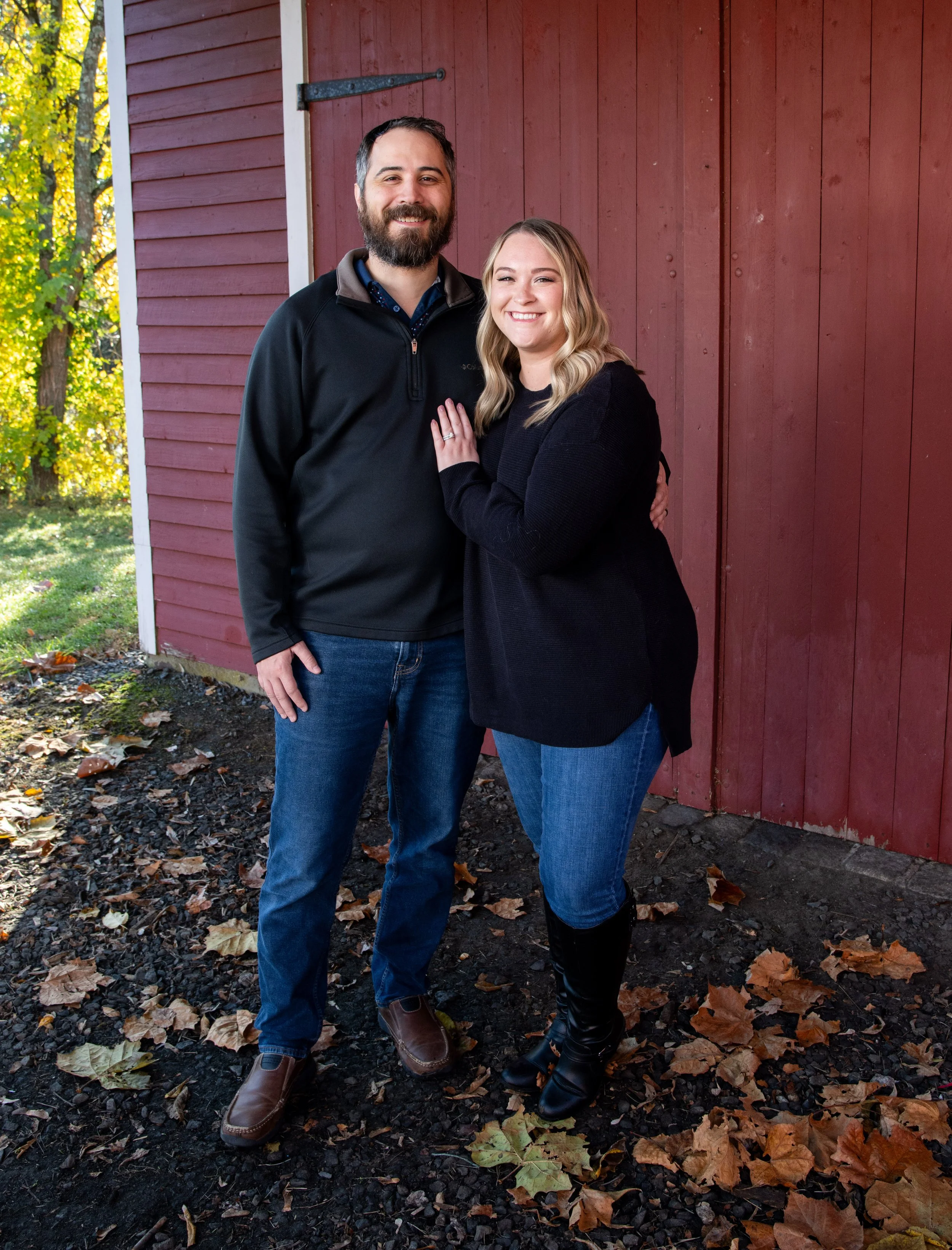 A smiling man with a beard and a woman with wavy blonde hair pose outdoors beside a red wooden building with trees and fallen leaves on the ground.