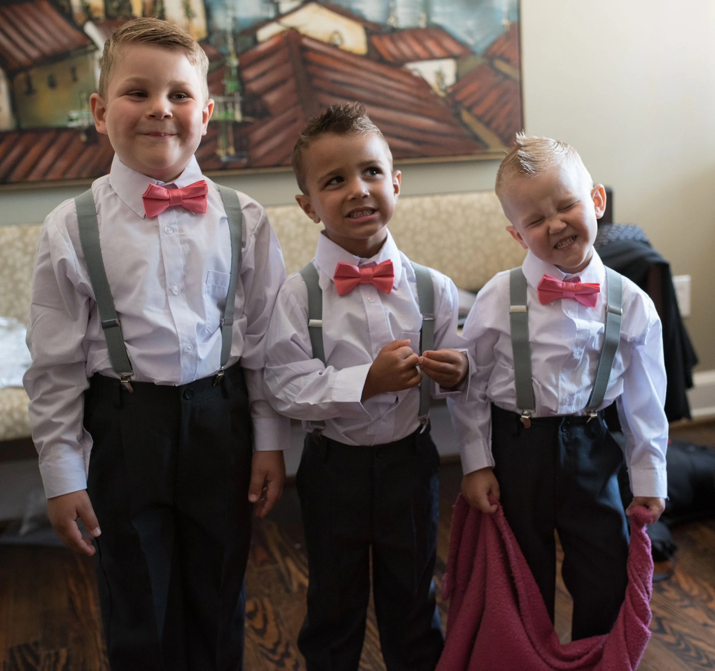 Three young boys dressed in formal attire with white shirts, pink bow ties, and suspenders, standing close together indoors. One boy is smiling, another is making a funny face, and the third is grimacing, with a colorful painting of rooftops in the b
