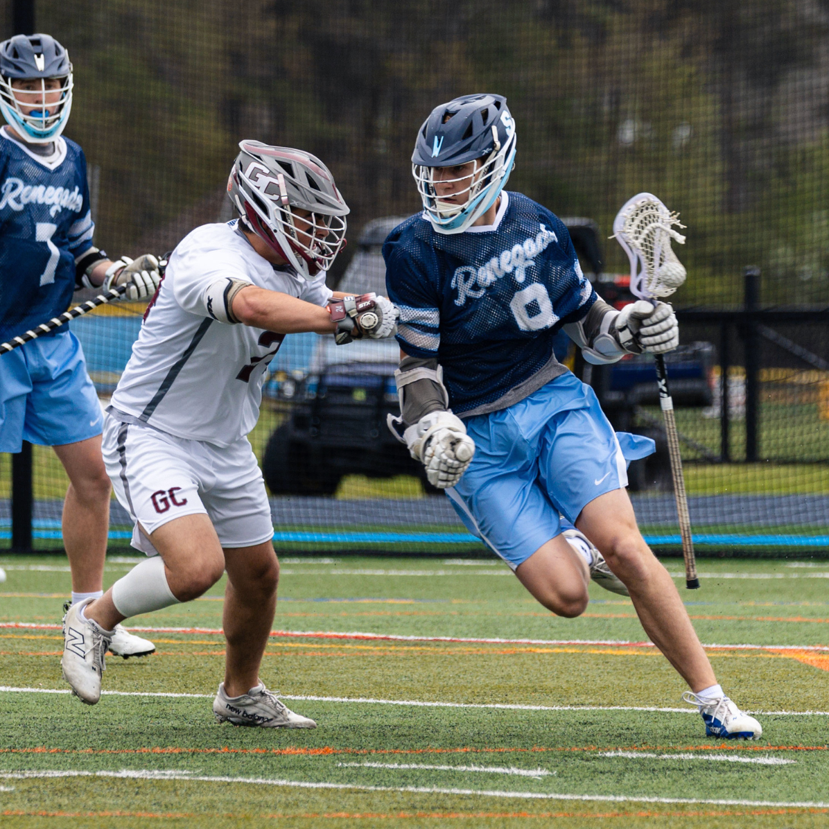 Lacrosse players in action on the field, with one player in a blue jersey and another in a white jersey competing for the ball.