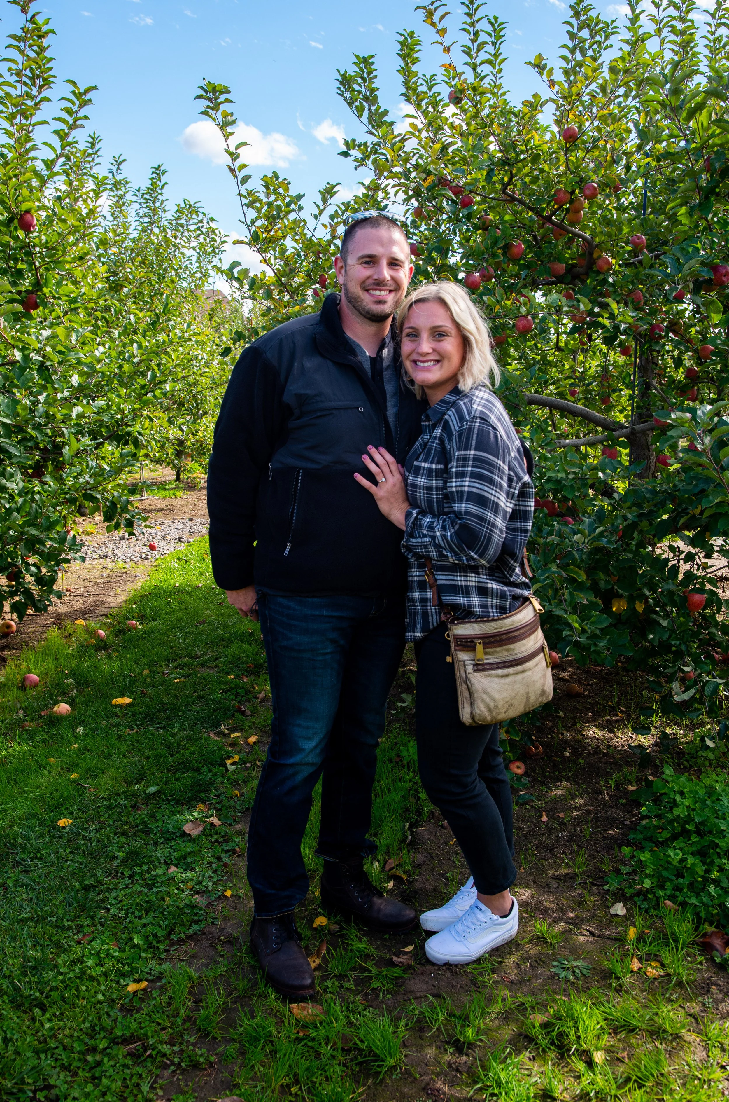 A happy couple stands together in an orchard with apple trees behind them. The man is wearing a black jacket and jeans, and the woman is wearing a plaid shirt, black pants, and white sneakers. They are smiling and holding each other, with a bright bl