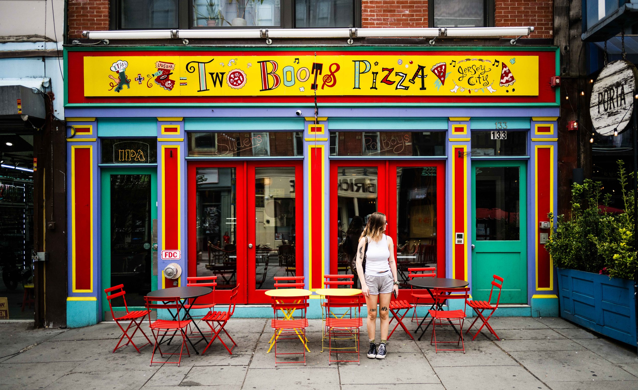 Colorful storefront of a pizza restaurant with yellow, red, and blue accents, outdoor seating with red and yellow chairs and black tables, and a woman standing outside wearing a white shirt, grey shorts, and black sneakers.