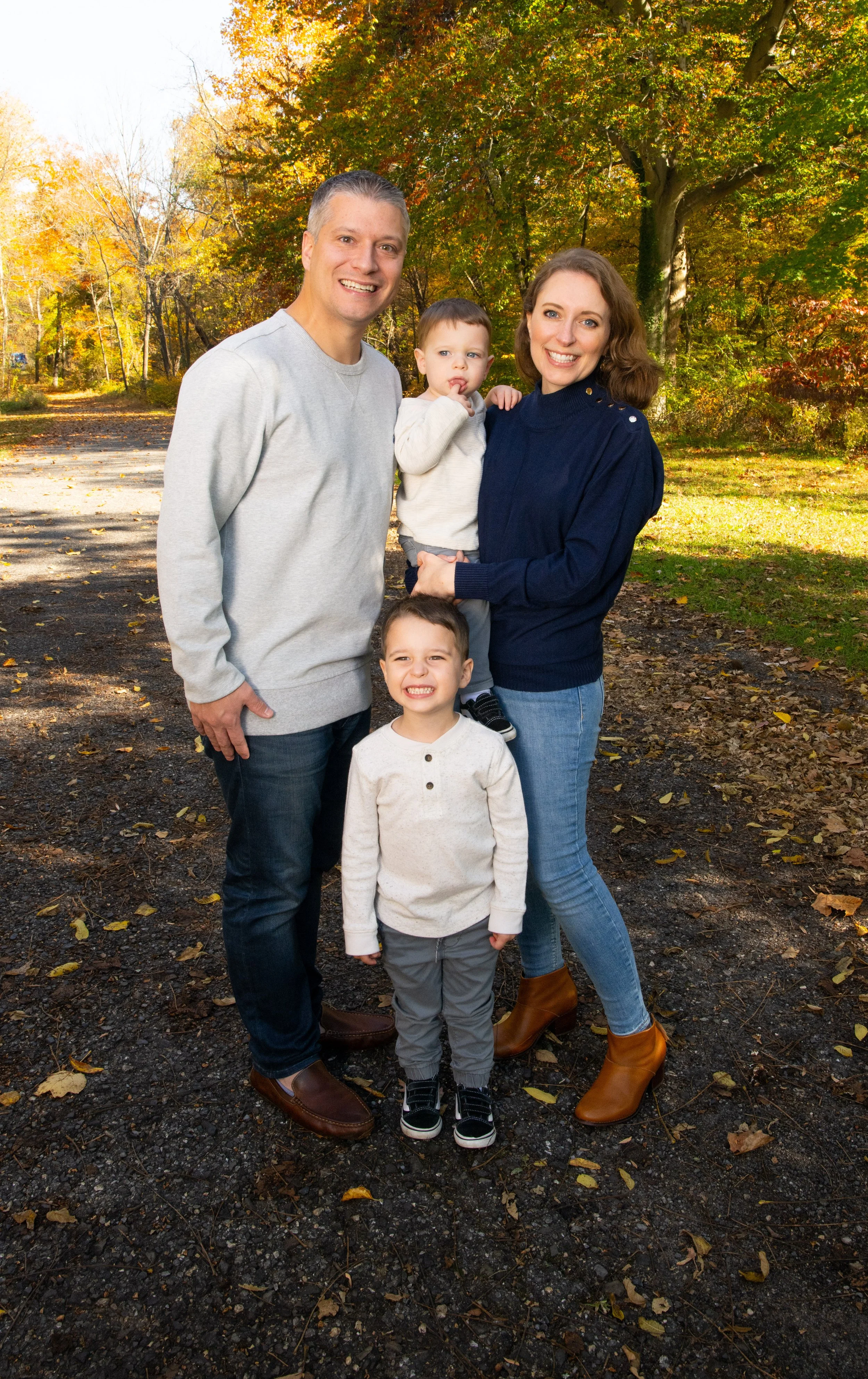 A family of five standing outdoors among trees with fall foliage. The father, mother, and three young boys are smiling at the camera. The father is wearing a light gray sweater, and the mother is in a dark blue sweater and brown boots. The children a