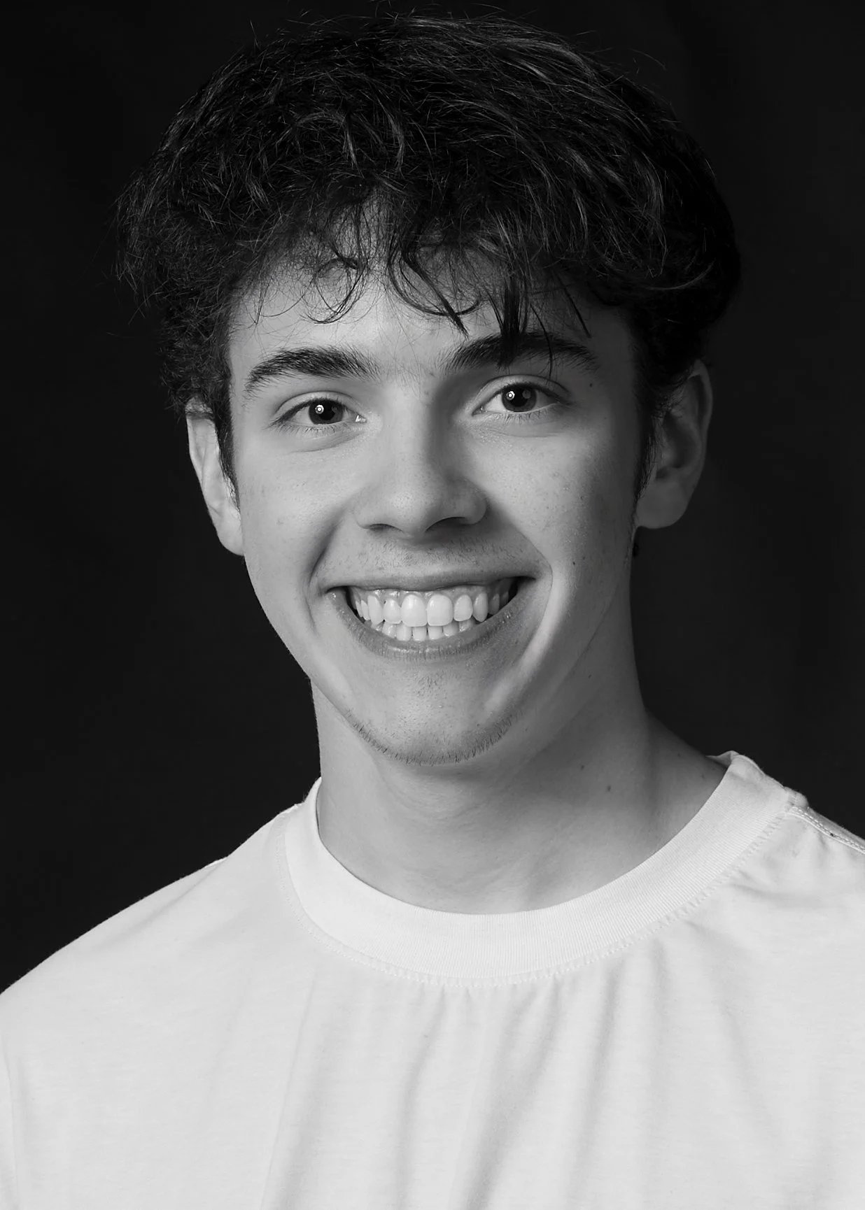 Black and white portrait of a young man with curly hair, smiling, wearing a white t-shirt, against a dark background.