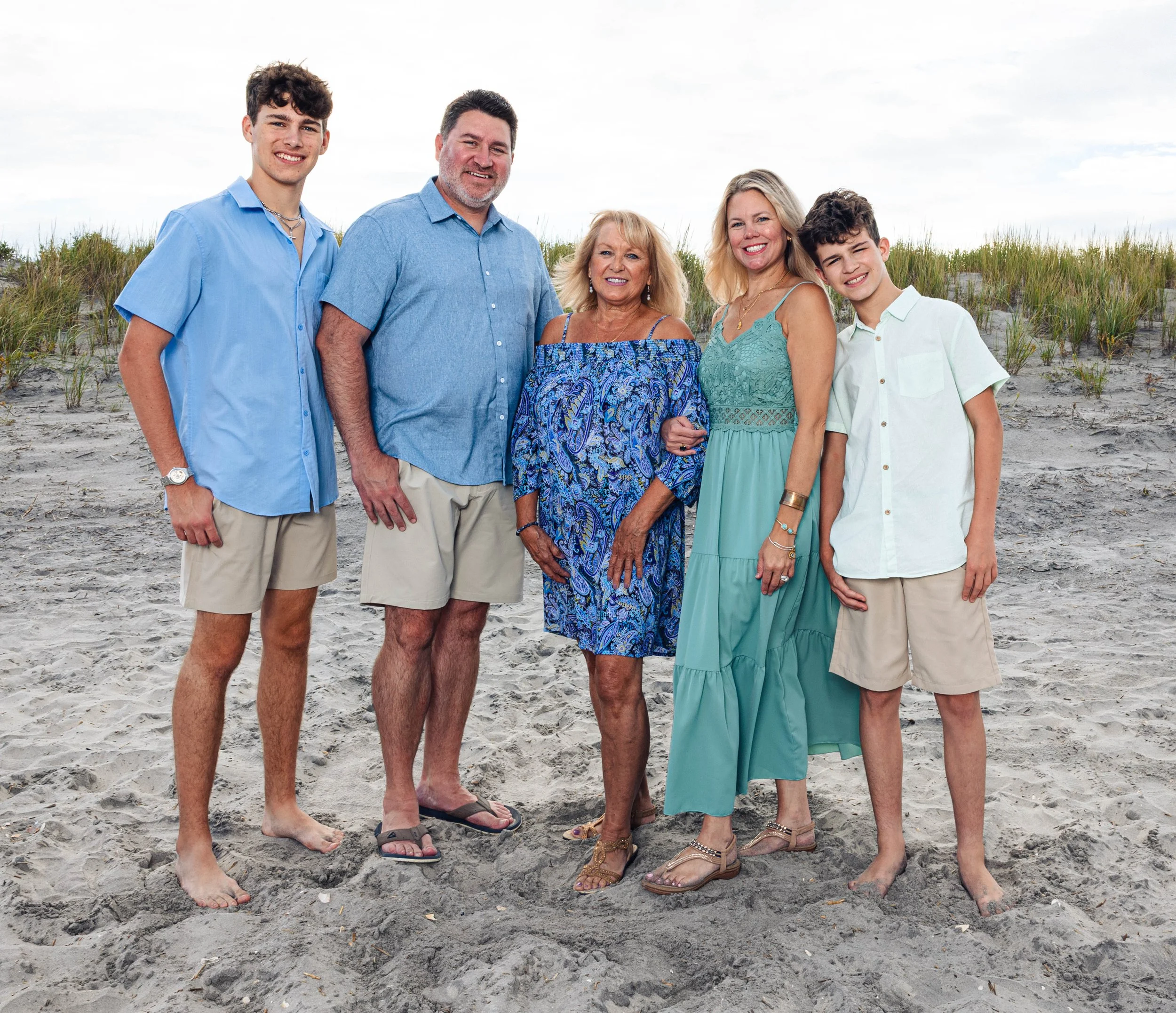 A multi-generational family standing on a sandy beach with grassy dunes in the background, smiling at the camera.