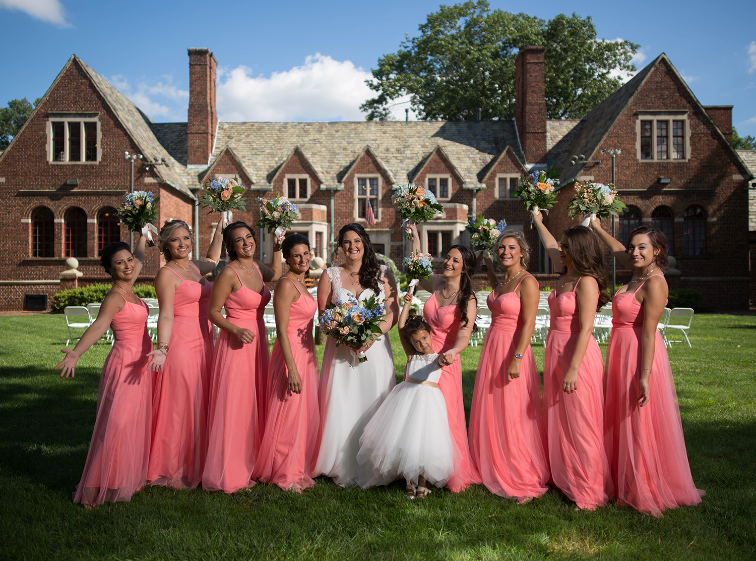 A bride and ten bridesmaids standing outside on a lawn in front of a large brick mansion under a blue sky. The bride is in a white wedding dress holding a bouquet, and the bridesmaids are in pink dresses, some holding bouquets with arms raised. A you