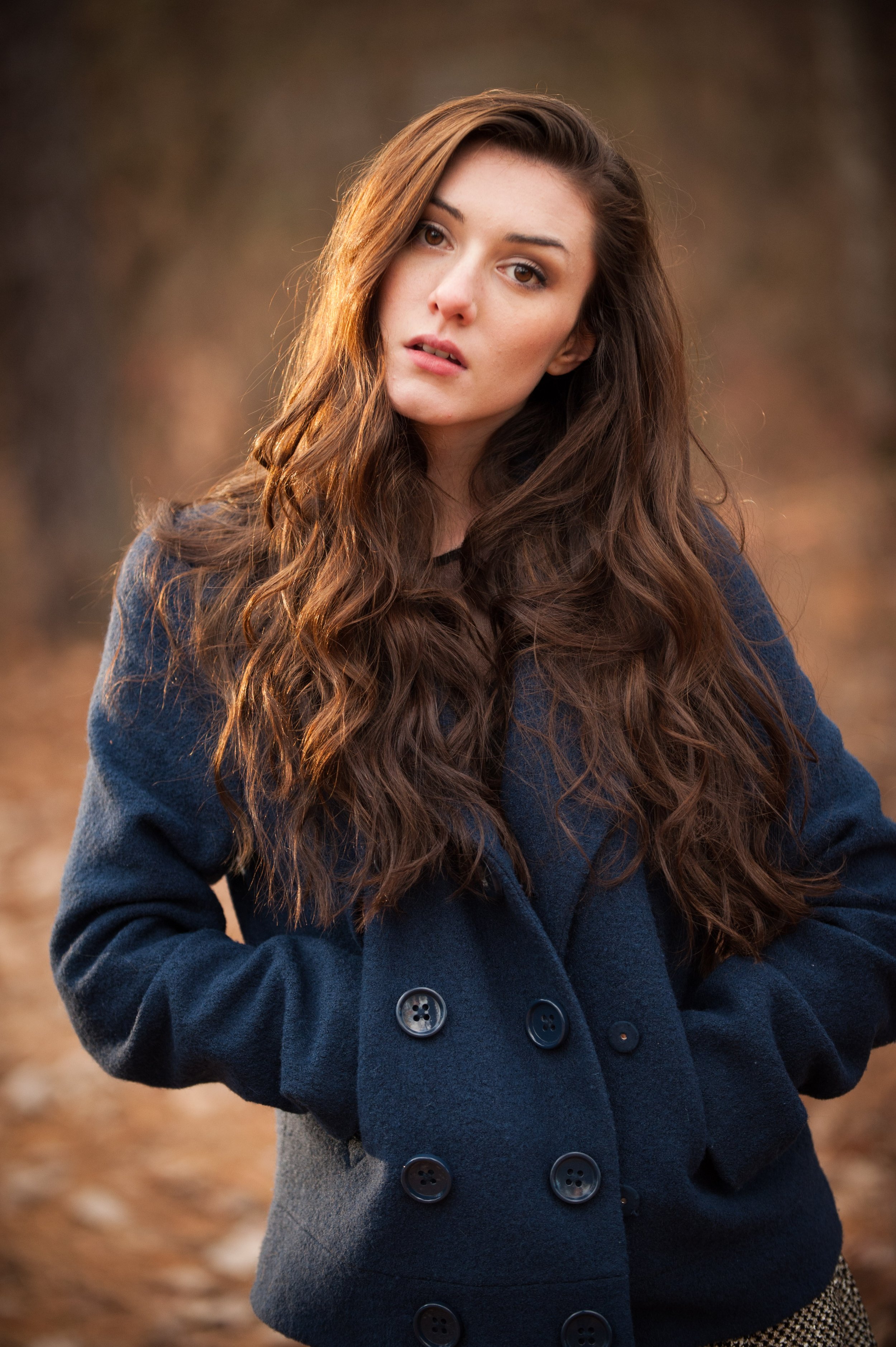 A young woman with long wavy brown hair, light makeup, and an expression with slightly parted lips, wearing a navy blue coat with large buttons, outdoors with blurred earthy background.