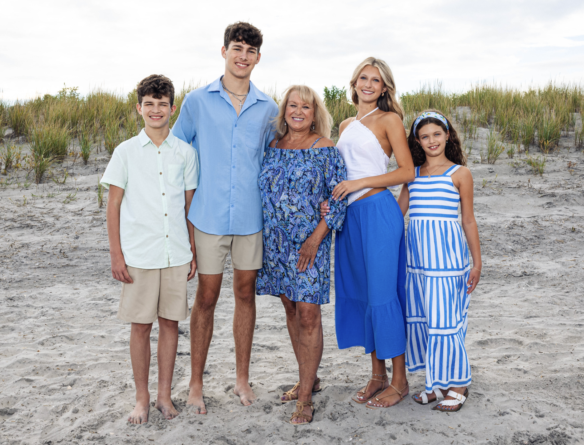 A group of six family members standing on a sandy beach with grass in the background, smiling for the camera, dressed in summer clothing.