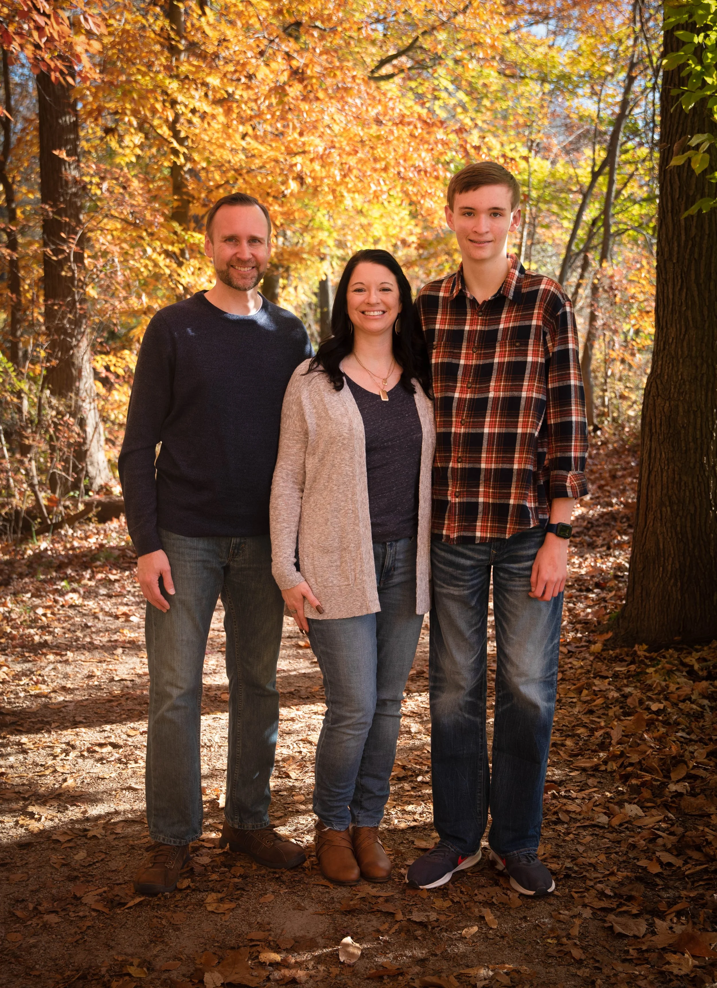 A smiling family of three standing in an autumn park surrounded by orange and yellow leaves.
