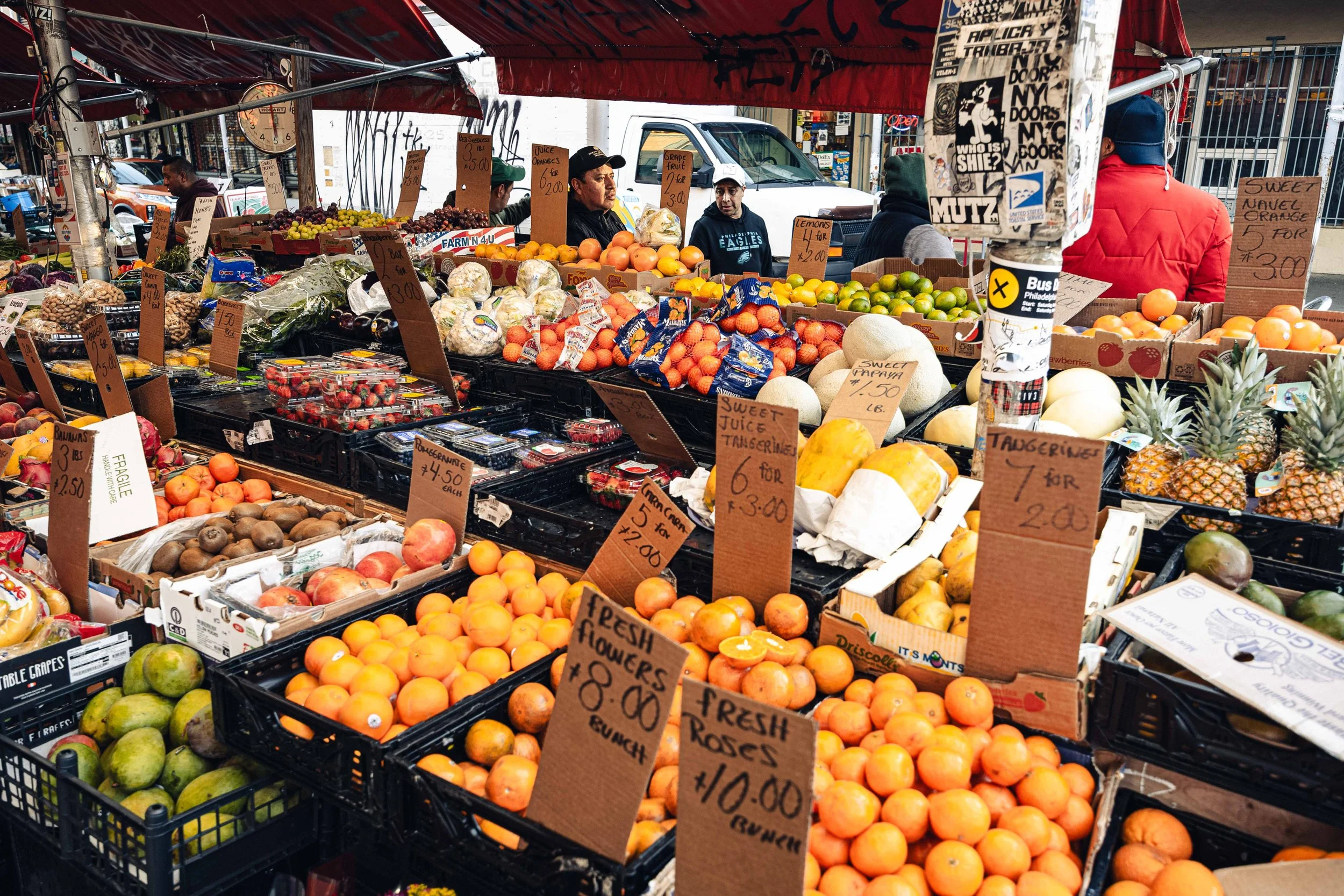Market stall with fresh fruits and vegetables, including oranges, peaches, strawberries, kiwis, pineapples, and cauliflower, with handwritten price signs. Several vendors and customers are visible in the background under a red canopy.