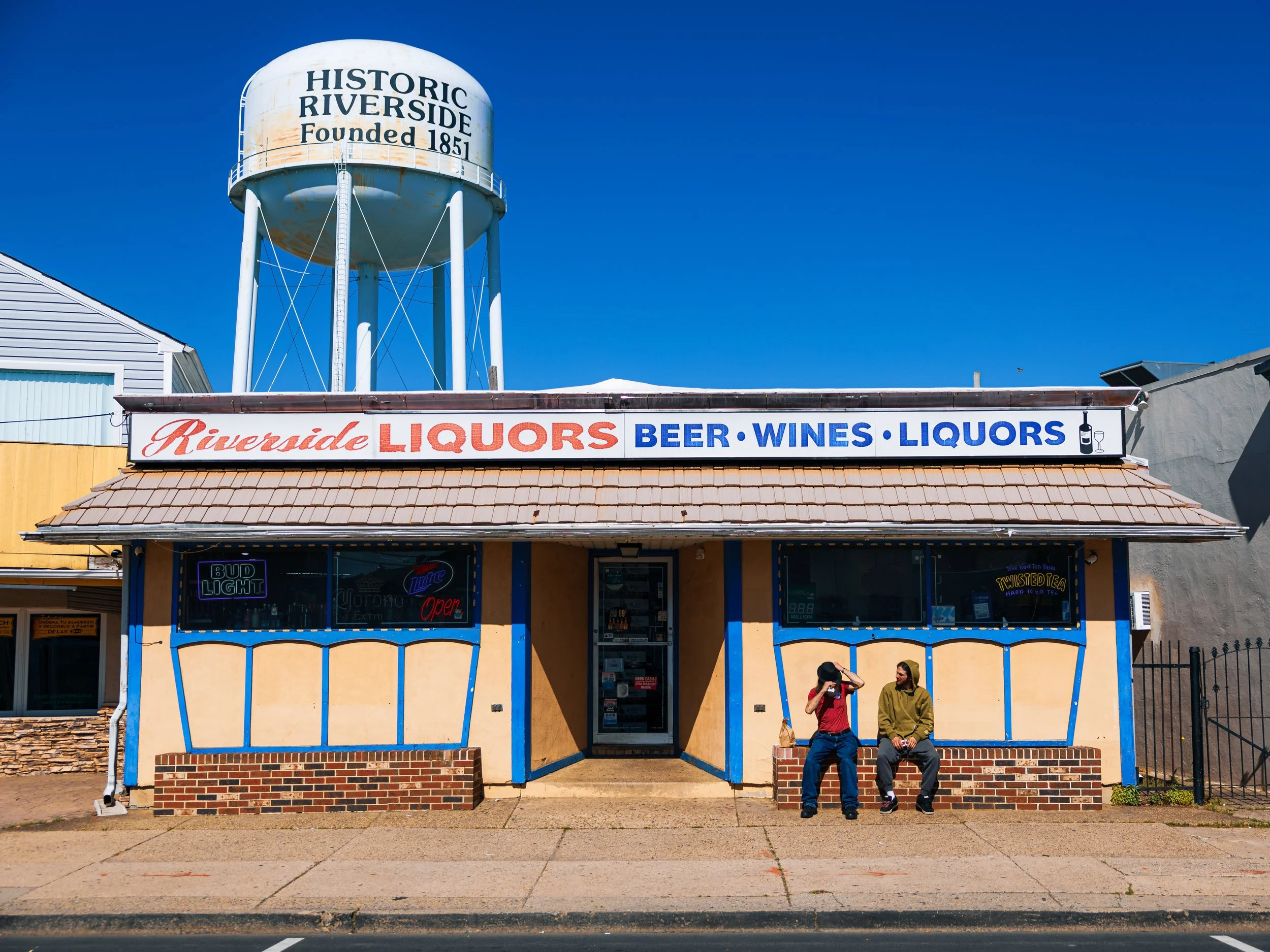 A building with a sign reading 'Riverside Liquors Beer Wines Liquors' in large red, blue, and black letters on the storefront, two people sitting on the sidewalk in front, and a water tower in the background with the words 'Historic Riverside Founded