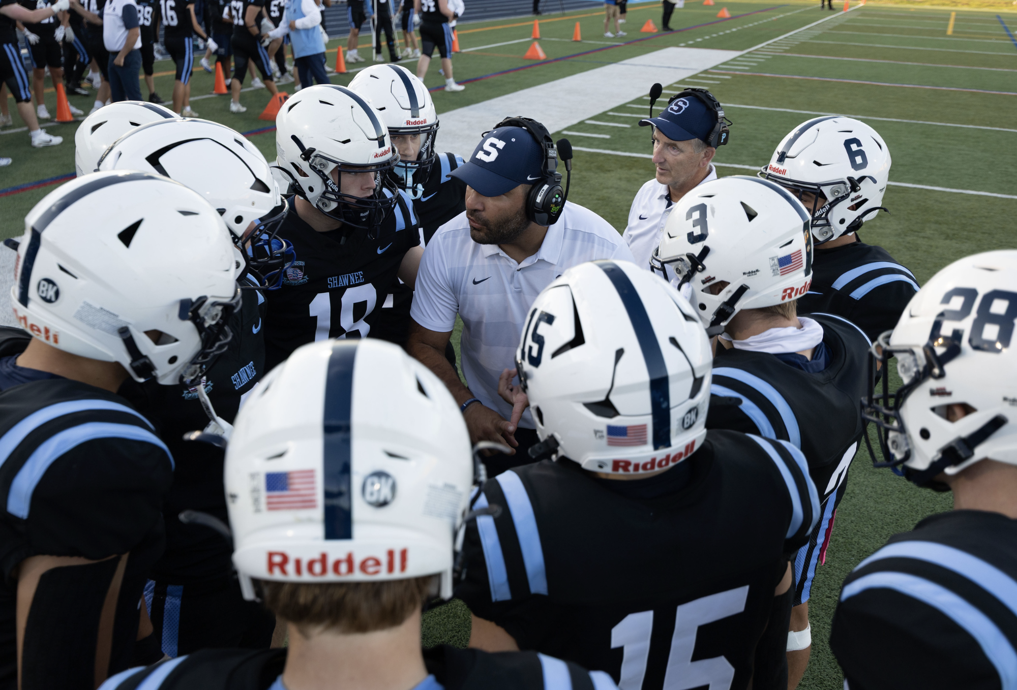 Football coach giving instructions to a team of young players during a timeout on the field. The players are wearing helmets and black jerseys with white helmets and blue accents. The team is huddled together closely as they listen to the coach.