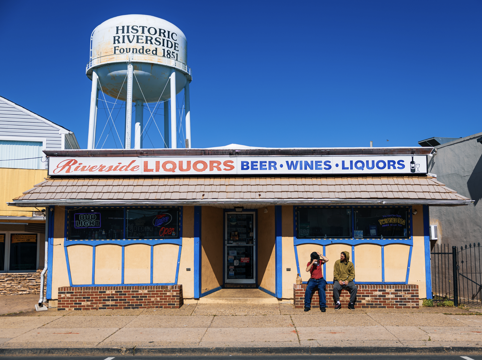 A storefront with a sign reading 'Riverside Liquors Beer Wines Liquors' and a neon 'Open' sign in the window. Above the store, a large water tower says 'Historic Riverside Founded 1854' against a clear blue sky. Two people are sitting on the sidewalk