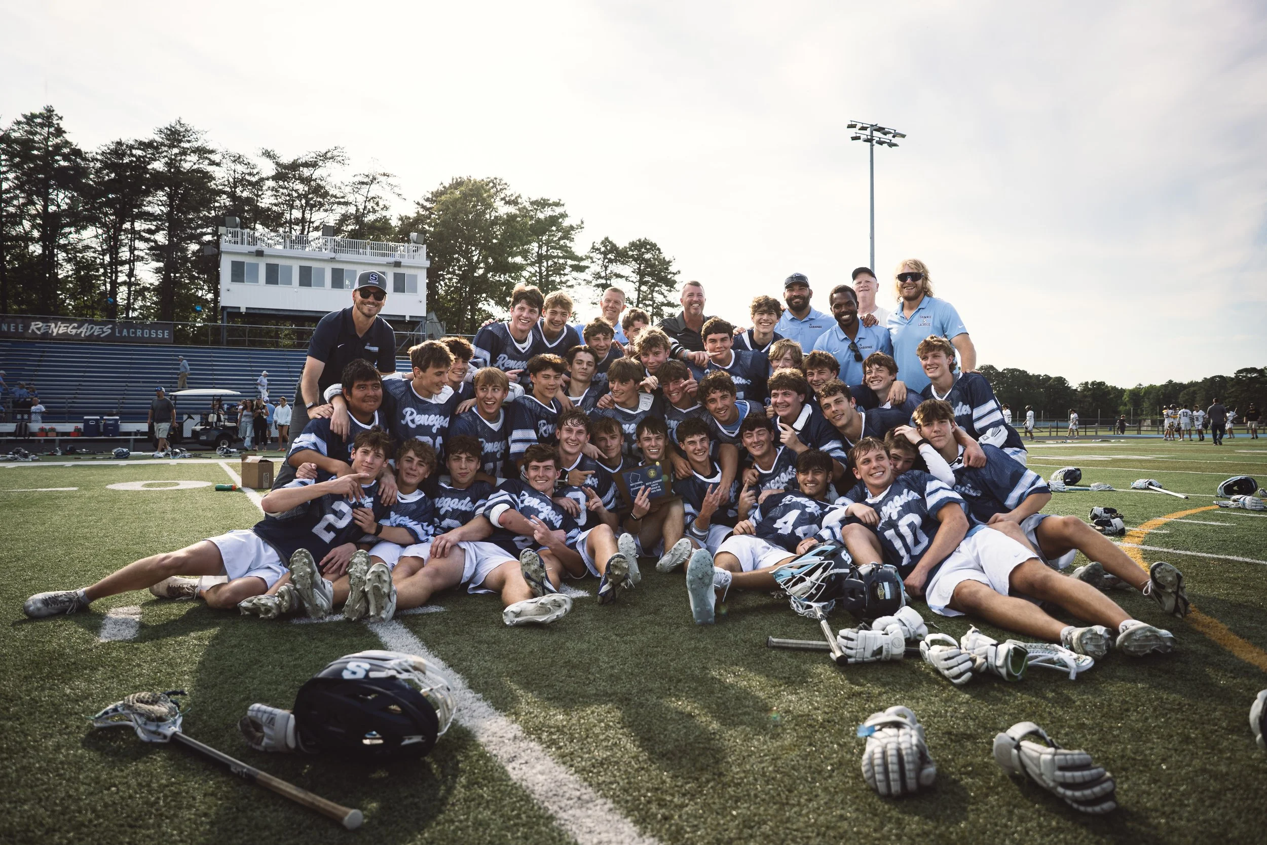 A group of young lacrosse players and their coaches posing for a team photo on a sports field after a game, with lacrosse equipment in front.