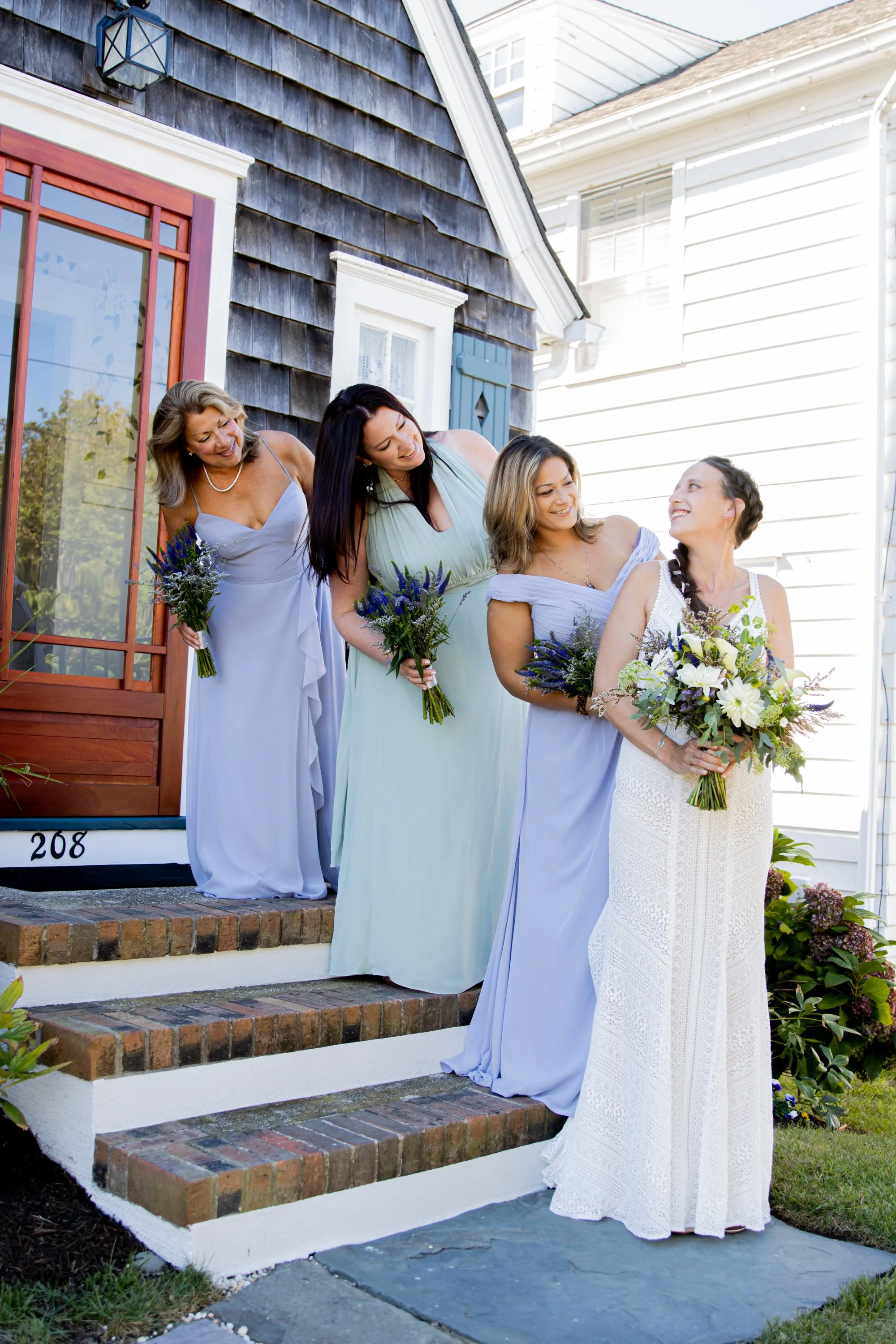 Four women dressed in formal light-colored gowns are standing on the steps outside a house, holding bouquets of flowers and smiling at each other.