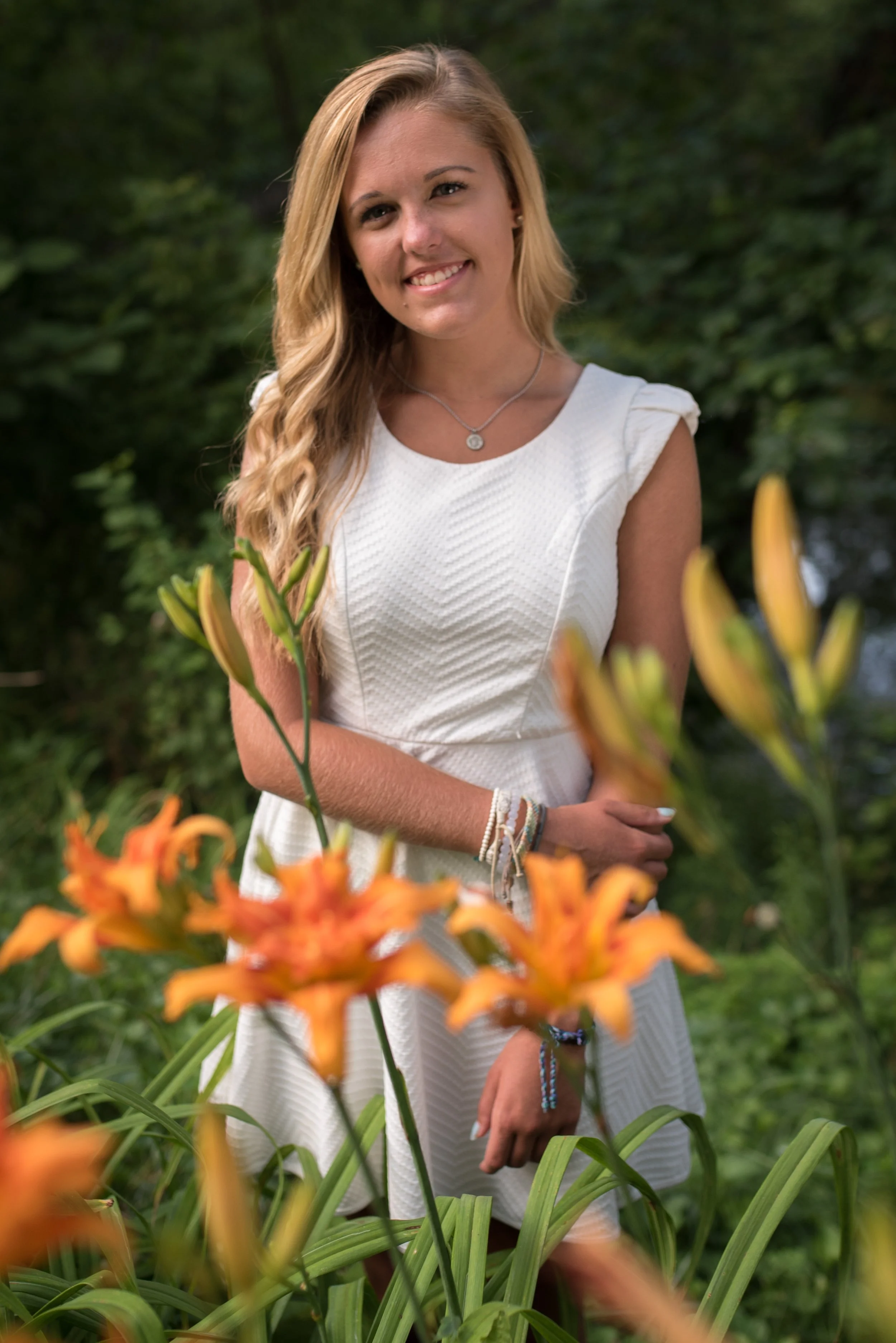 A woman with long blonde hair, smiling, is standing outdoors among orange daylilies. She is wearing a white sleeveless dress with a textured pattern, a necklace, and bracelets. The background is green foliage.