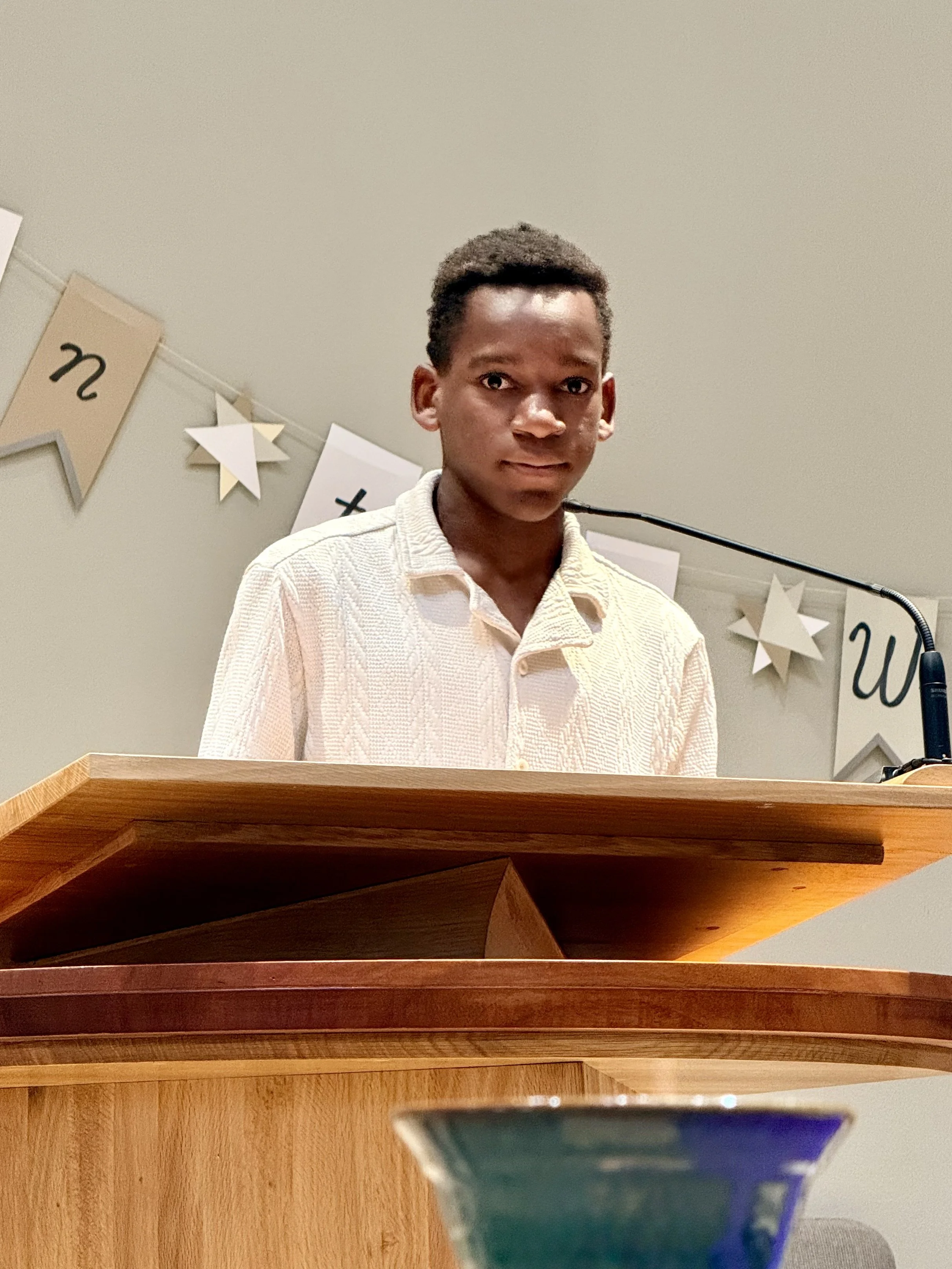 A young man standing behind a wooden desk with a microphone, wearing a light-colored textured shirt, in front of a wall with decorative paper banners and stars.