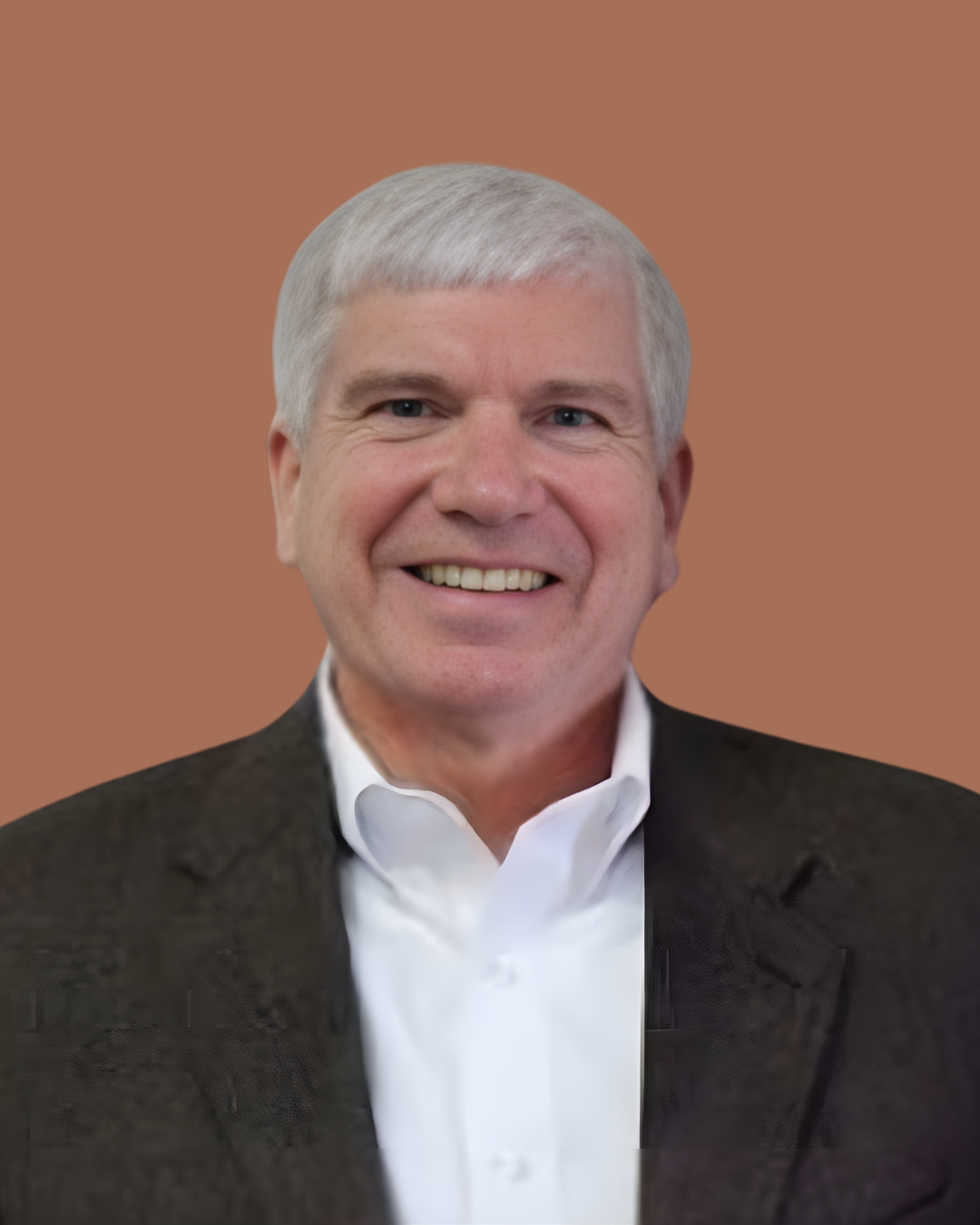 Headshot of a smiling middle-aged man with gray hair, wearing a white shirt and dark blazer, against a brown background.