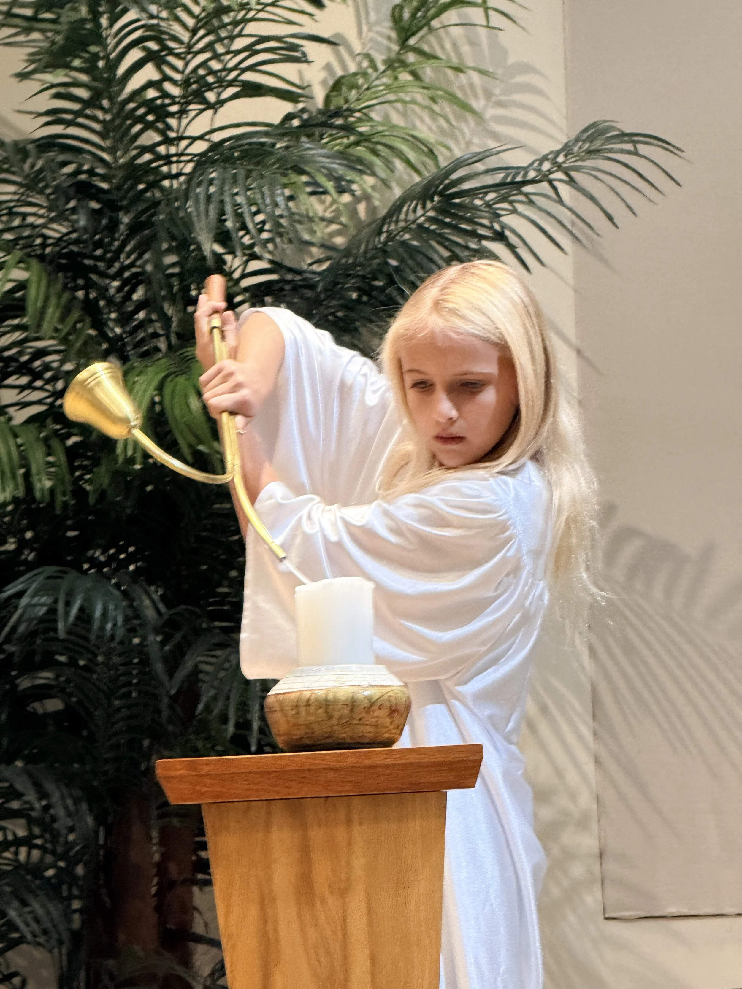 A young girl dressed in a white robe is lighting a white candle with a tall yellow candle, standing behind a wooden candle holder, with large green tropical plants in the background.