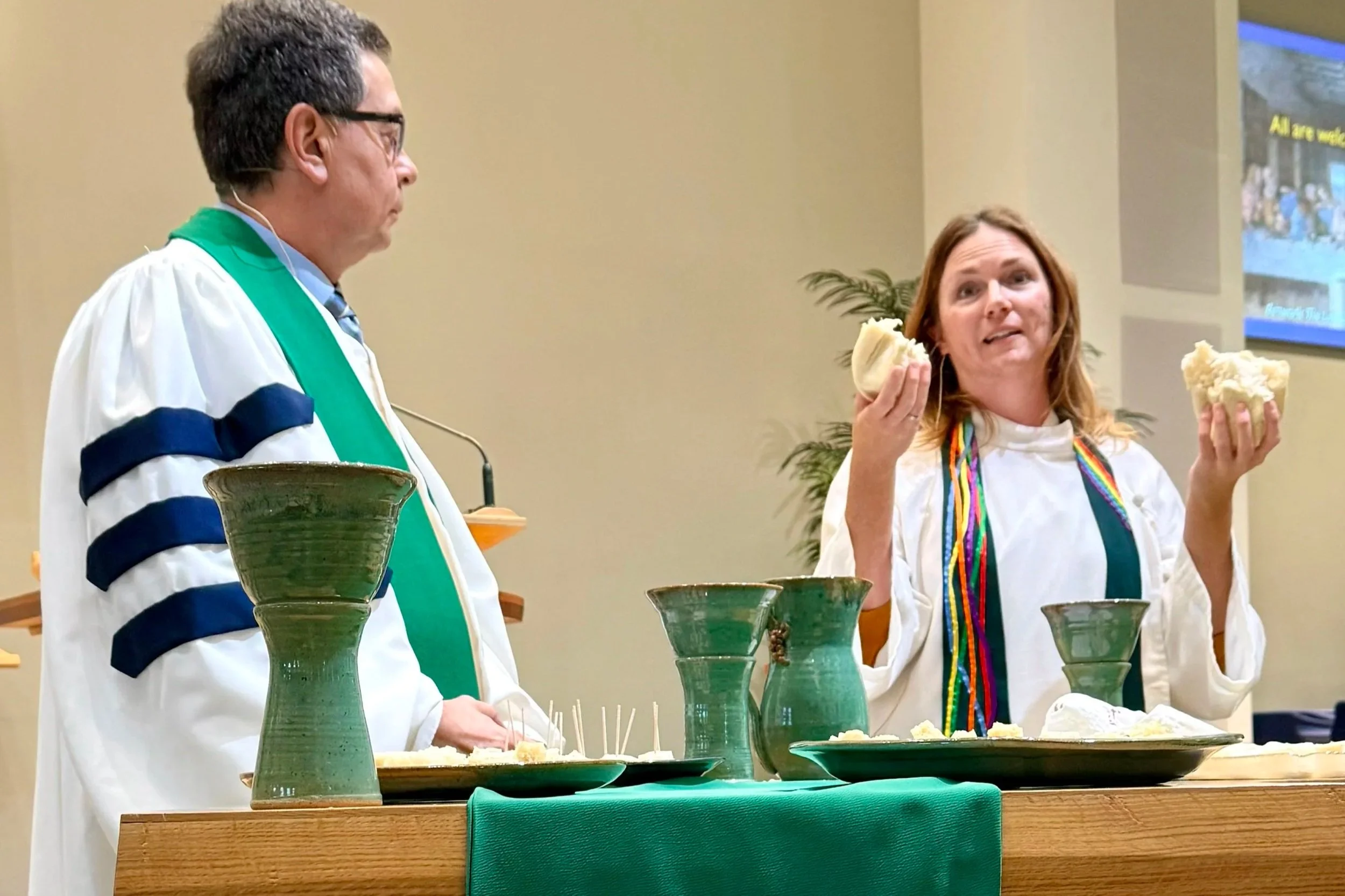 Two people in white robes with green stoles, standing behind a table set with communion elements, engaging in a conversation. The woman on the right is holding up pieces of bread, while the man on the left is looking at her.