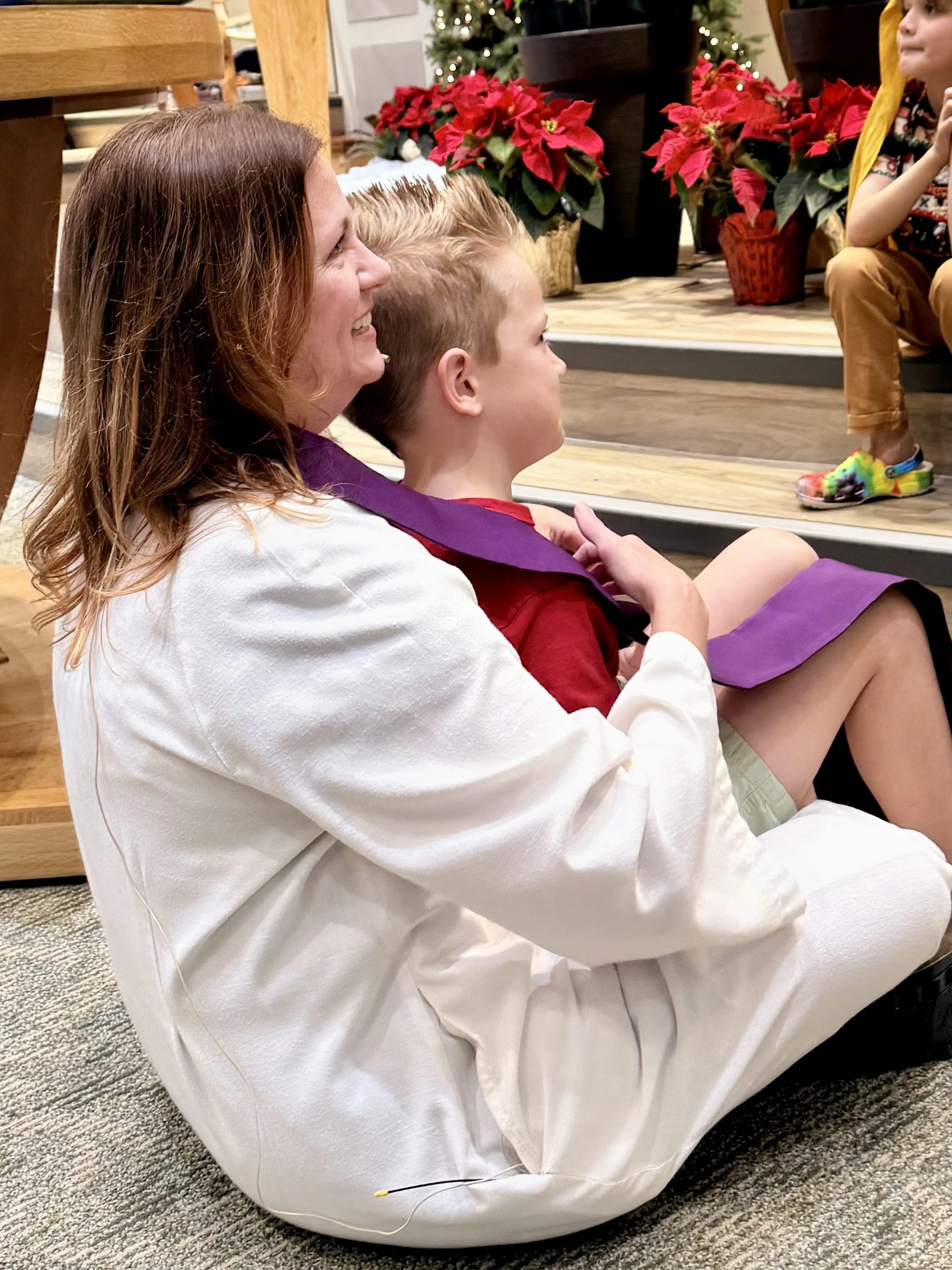 A woman and a young boy sitting on the floor, watching a performance or presentation. The woman is smiling, and the boy is wearing a purple scarf. Poinsettia plants and a decorated Christmas tree are visible in the background.