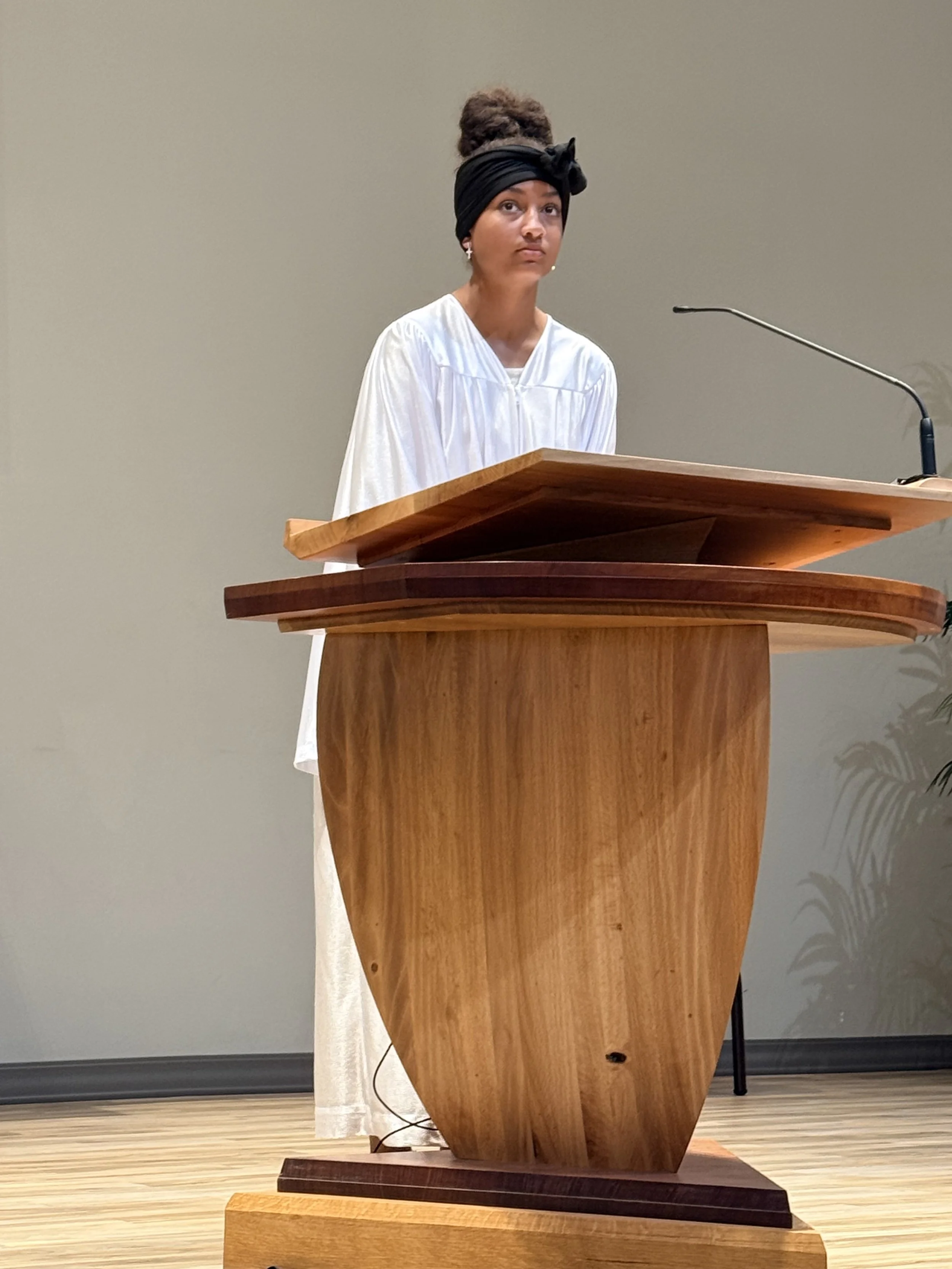 A woman with a black headwrap and white dress standing behind a wooden podium, looking serious, in a neutral-colored room.