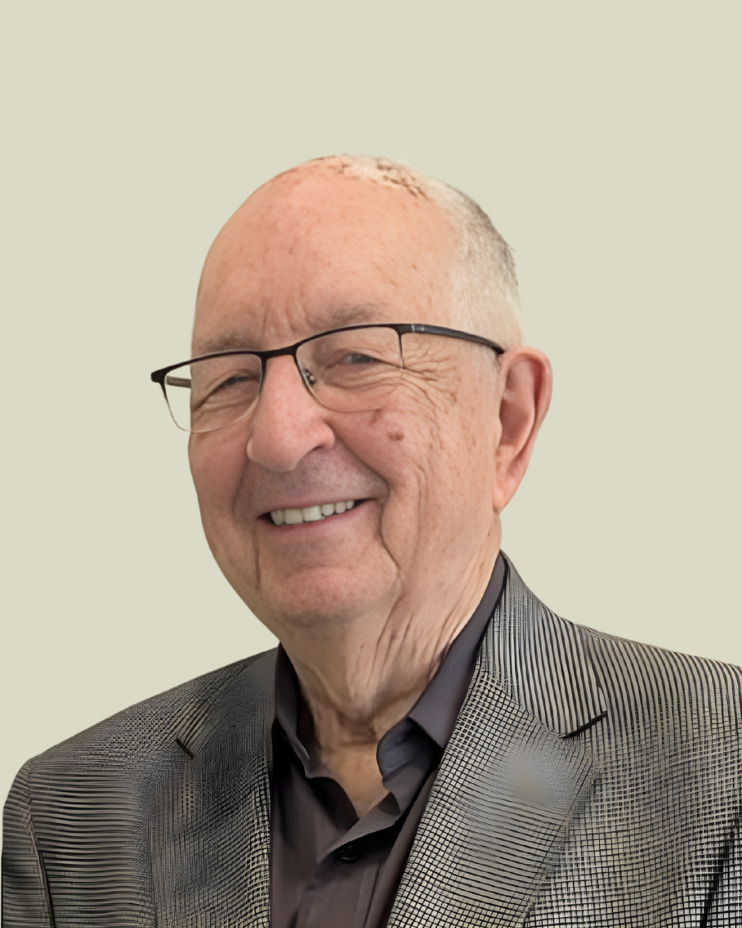 A smiling elderly man with glasses, wearing a checkered suit jacket and a dark shirt, standing against a plain light background.