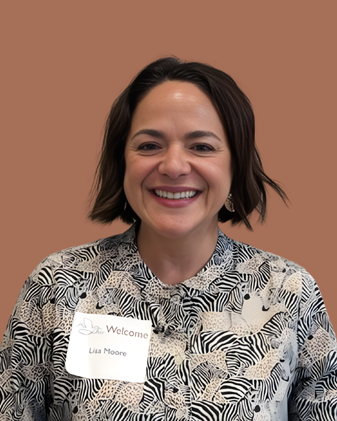 A smiling woman with shoulder-length dark hair wearing a patterned blouse with a name tag that says 'Welcome Lila Moore'.