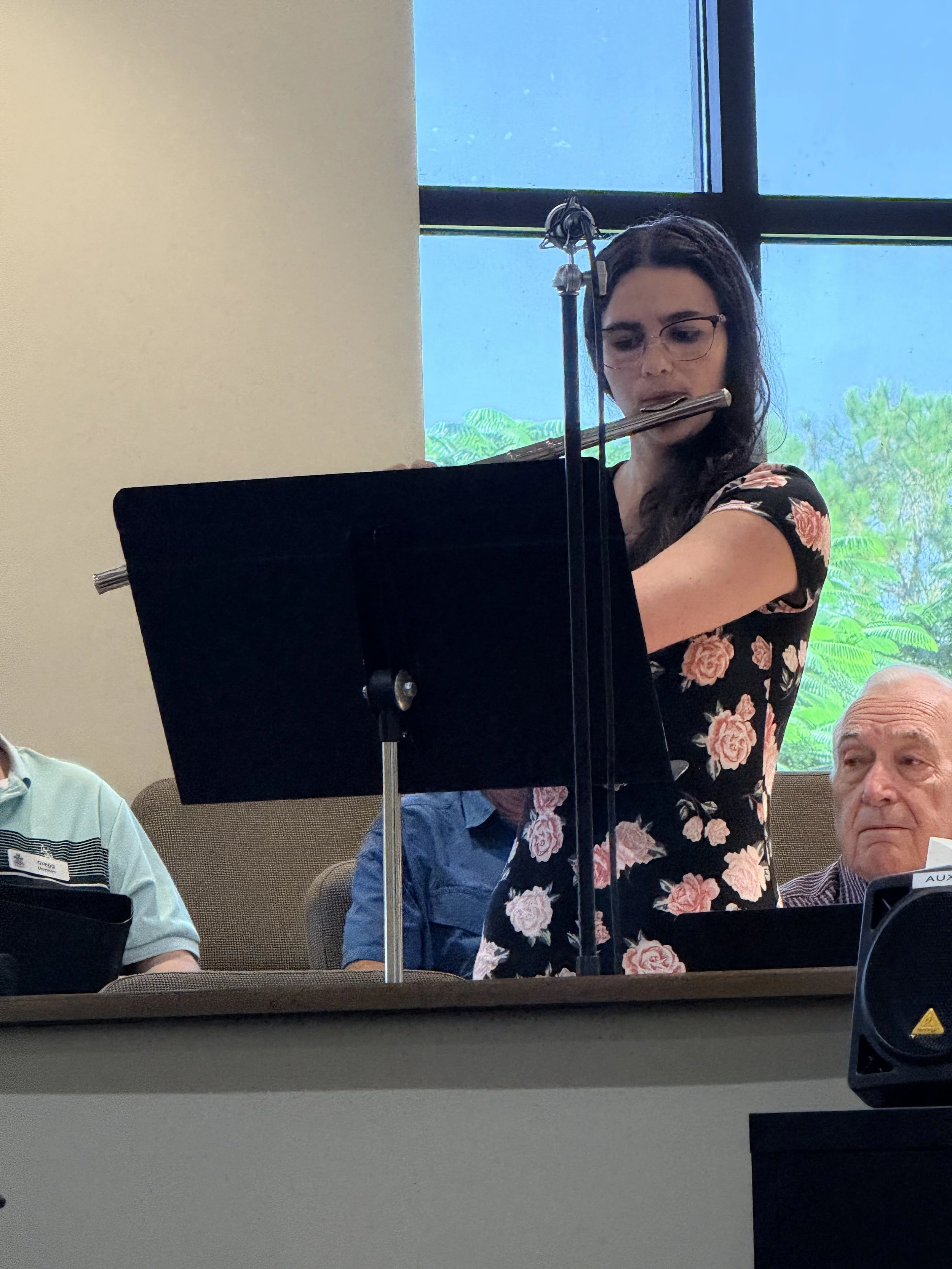A woman in a floral dress playing a flute during a musical performance in a room with a large window showing green foliage outside.