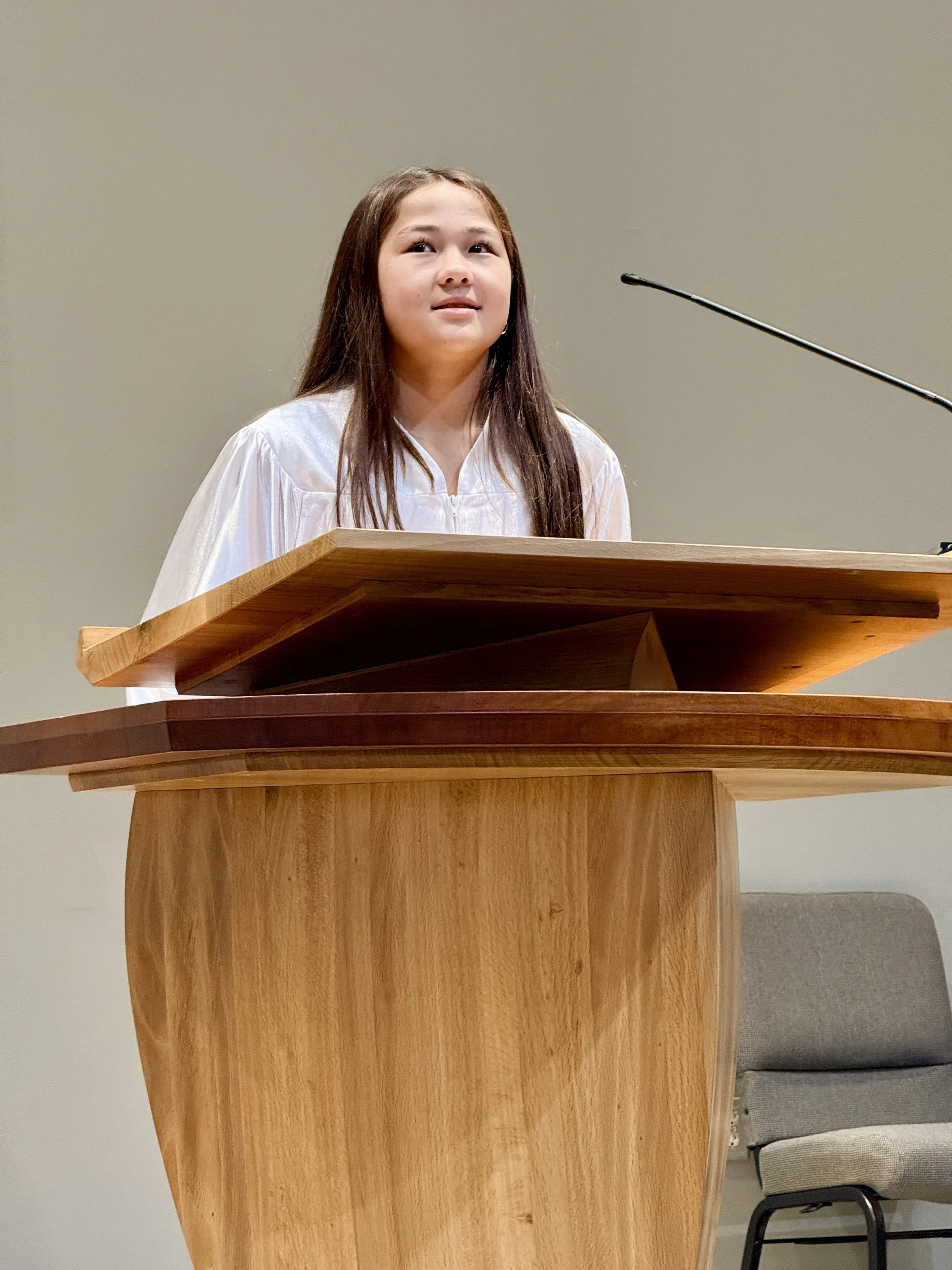 A young girl standing at a wooden podium, wearing a white gown, possibly giving a speech or presentation.