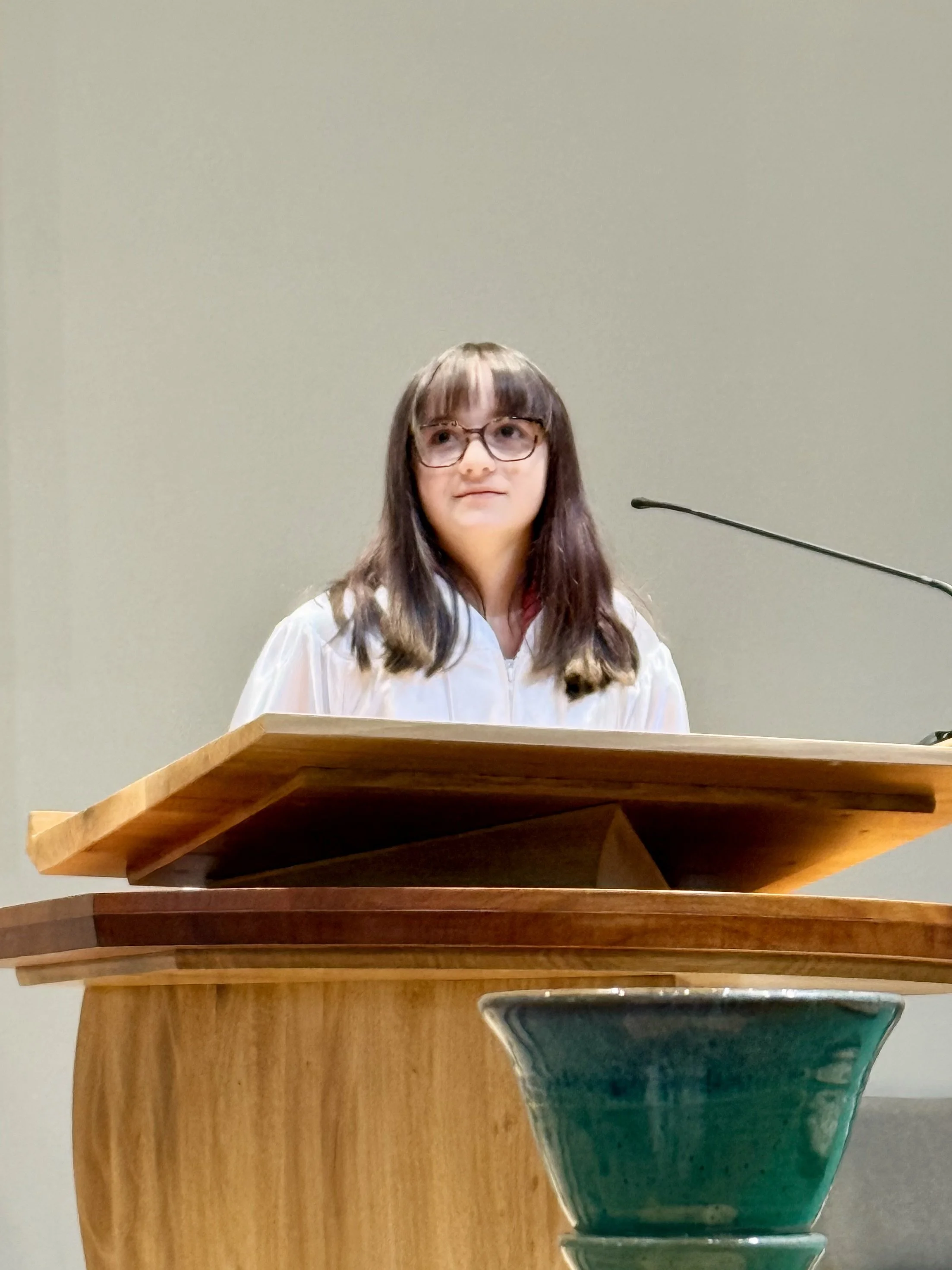 Young woman with glasses standing behind a wooden lectern, wearing a white graduation gown in a hall.