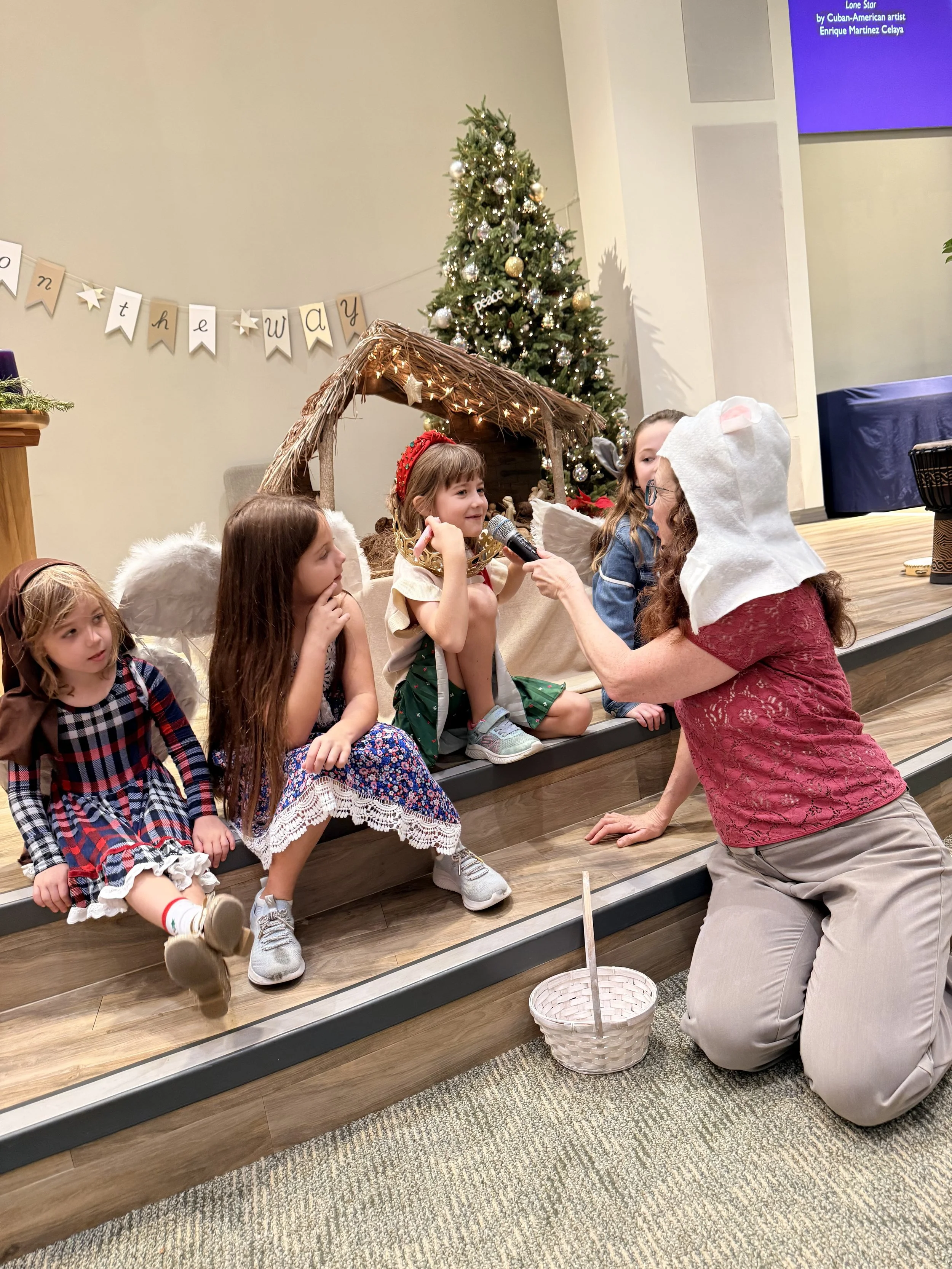 A woman wearing a white hood with ears and glasses is kneeling on the floor, holding a microphone for a young girl dressed as an angel. The girl is sitting on a platform with other children, all dressed in costumes, and a nativity scene with a Christ