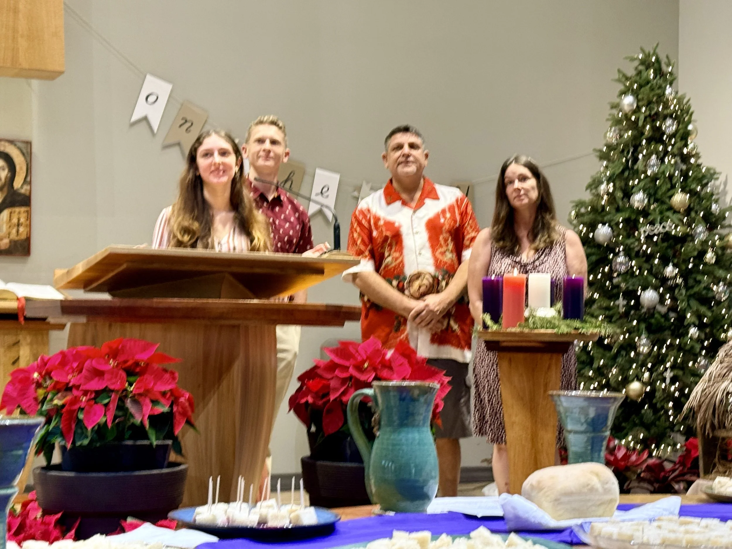Group of four people standing behind a table decorated with poinsettias and candles in a holiday setting with a Christmas tree in the background.