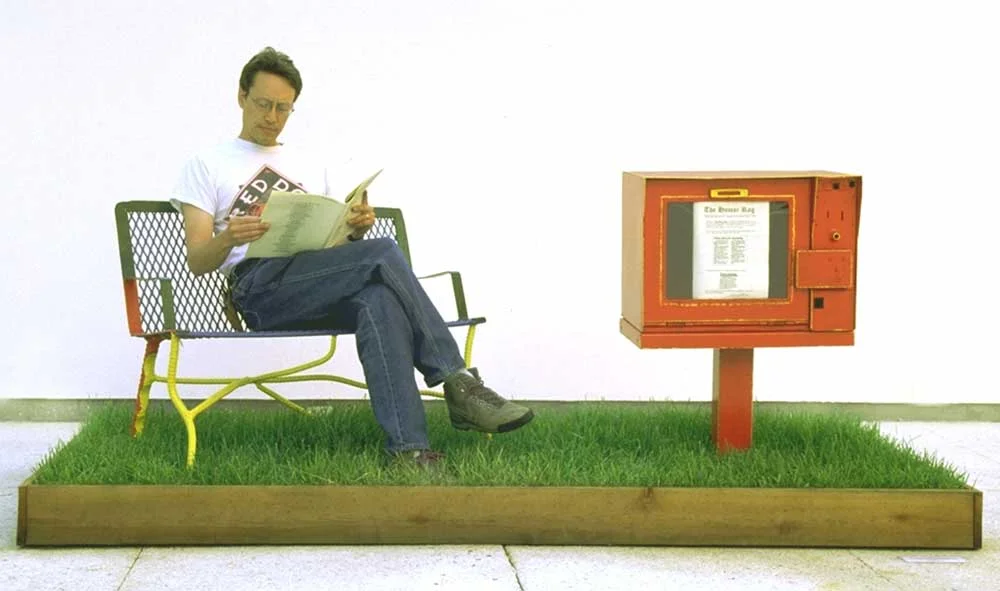 Humor Rag, an installation by Lorna Stevens featuring a newsstand holding a 40-page collection of jokes and witticisms, accompanied by grass, wood, and a bench.
