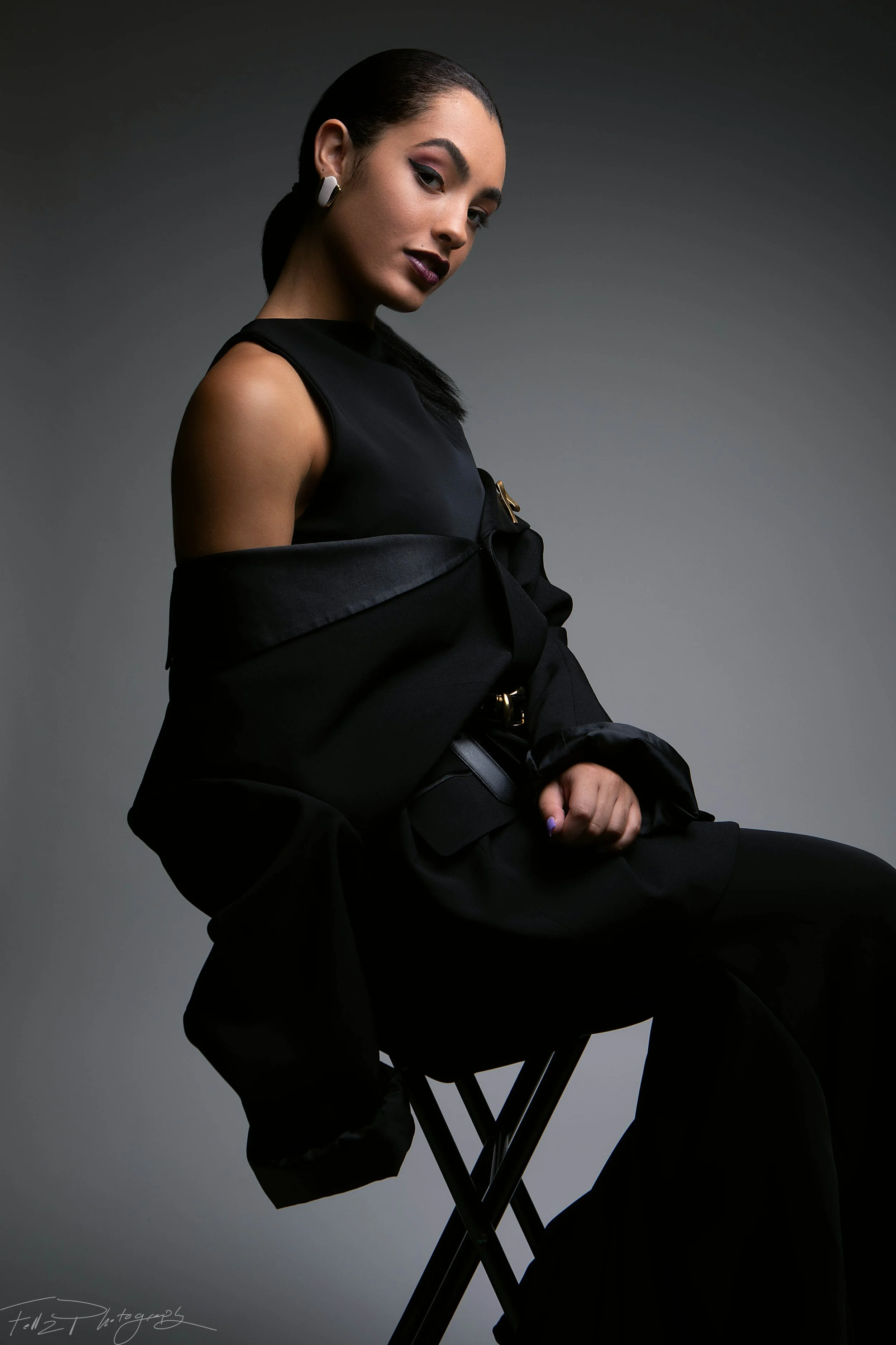 A woman with dark hair slicked back, wearing a sleeveless black top and matching black pants, sitting on a black folding chair against a grey gradient background, with a dark jacket draped over her shoulders.
