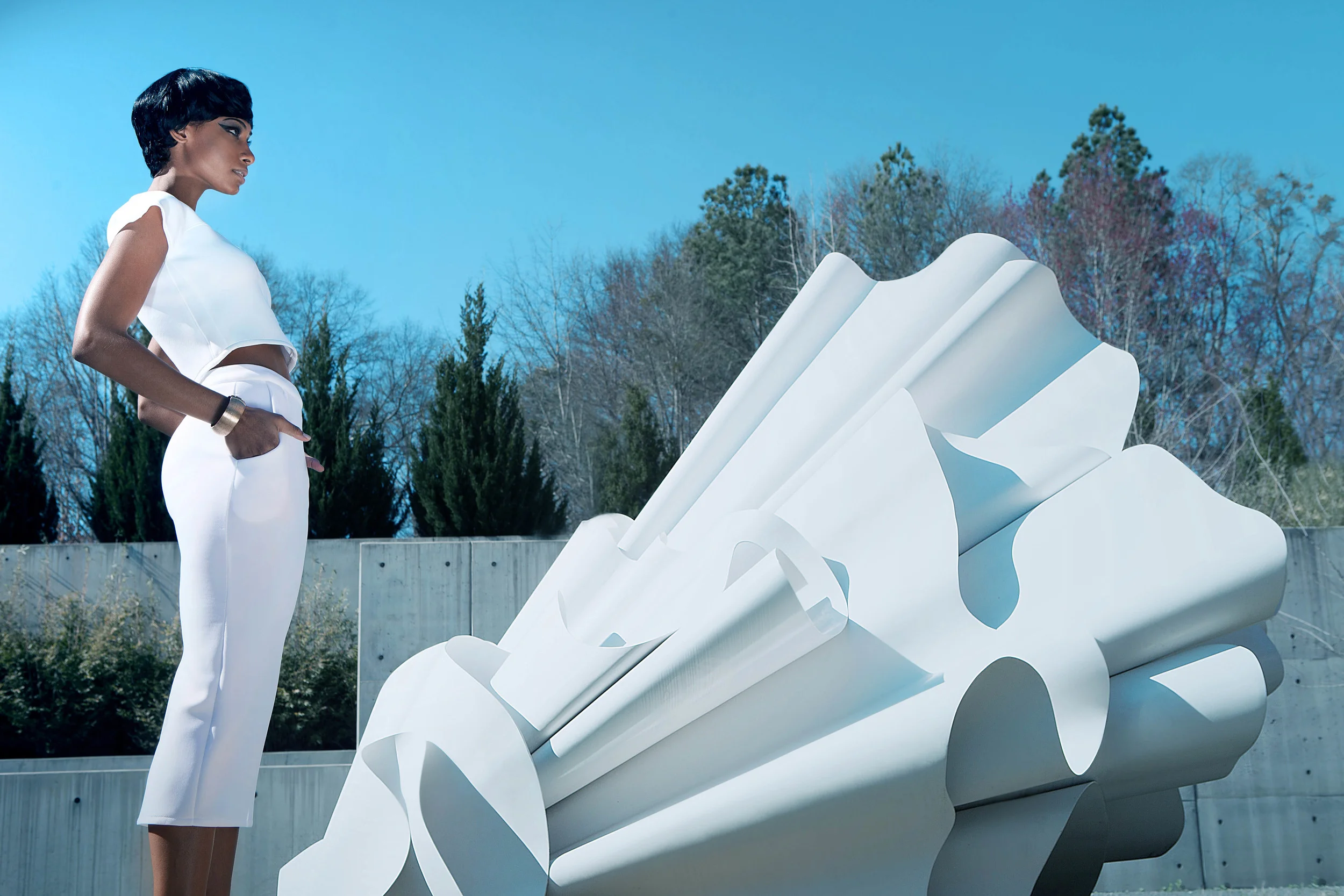 A woman in a white outfit stands outdoors with her hands in her pockets, looking at a large, abstract white sculpture set against a backdrop of trees and a blue sky.