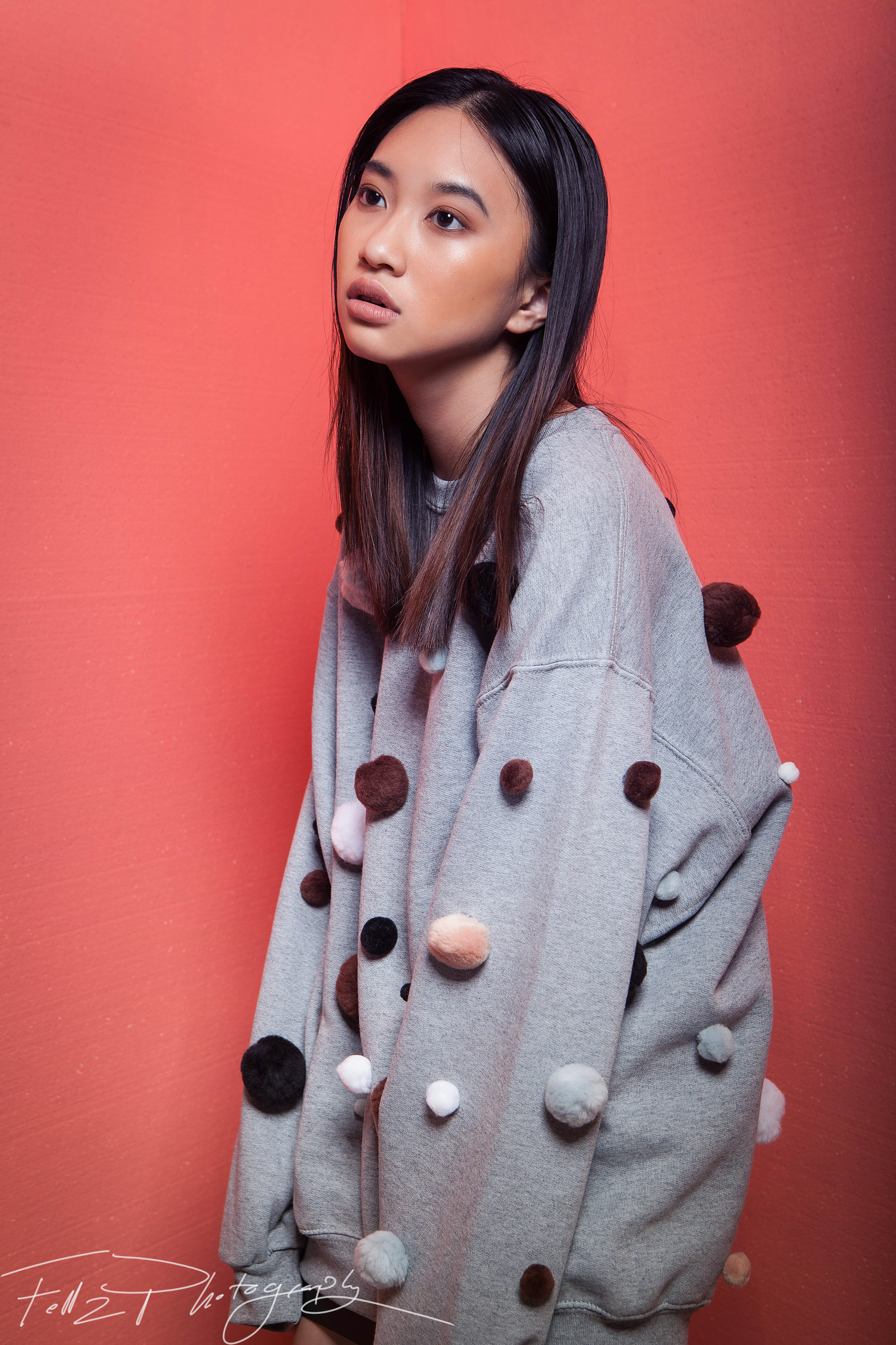 A young woman with straight black hair and light makeup stands against a pink wall. She wears a gray sweatshirt decorated with multicolored pom-poms.