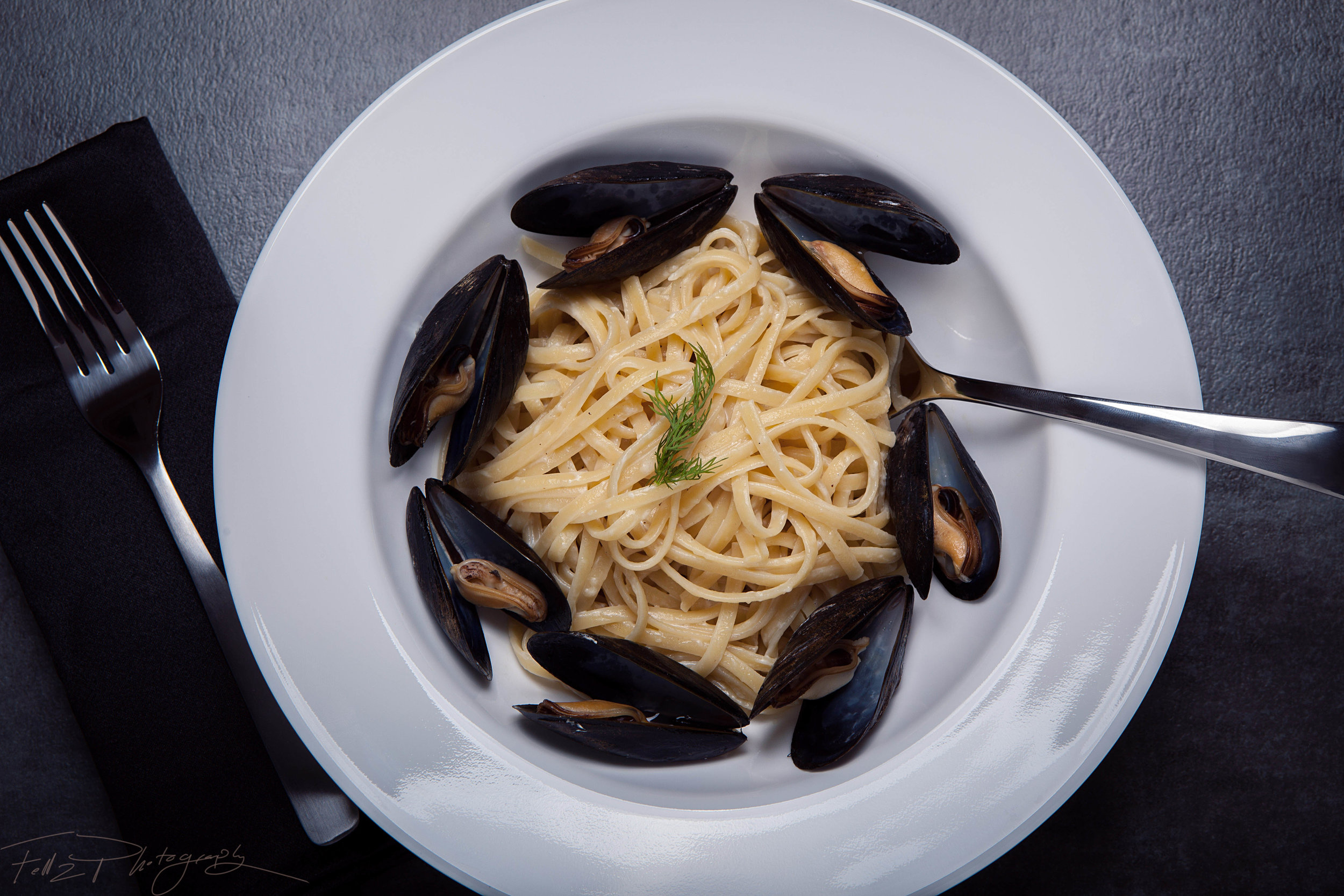 A plate of pasta with black mussels garnished with a sprig of dill, placed on a dark surface with a black napkin and fork to the left.