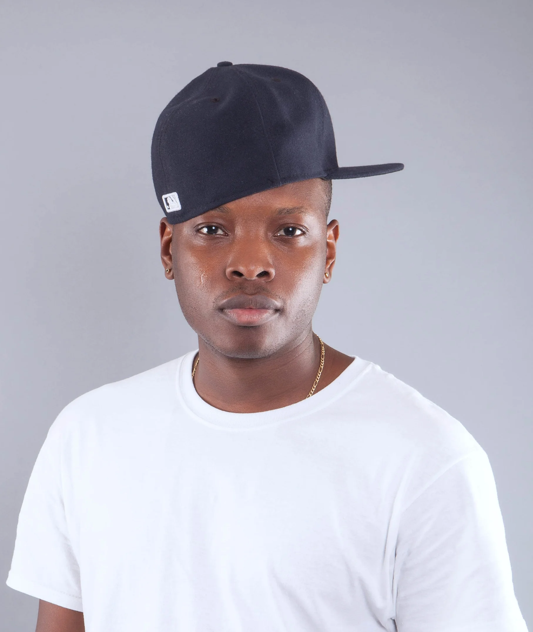A young man wearing a black baseball cap slightly tilted, a white t-shirt, gold earrings, and a delicate gold chain against a plain gray background.