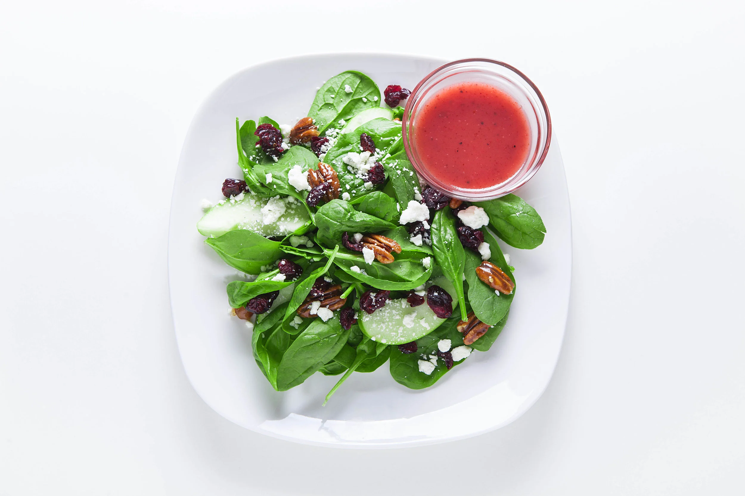 Fresh green spinach and mixed salad with dried cranberries, pecans, and crumbled cheese, served with a side of red salad dressing on a white plate.