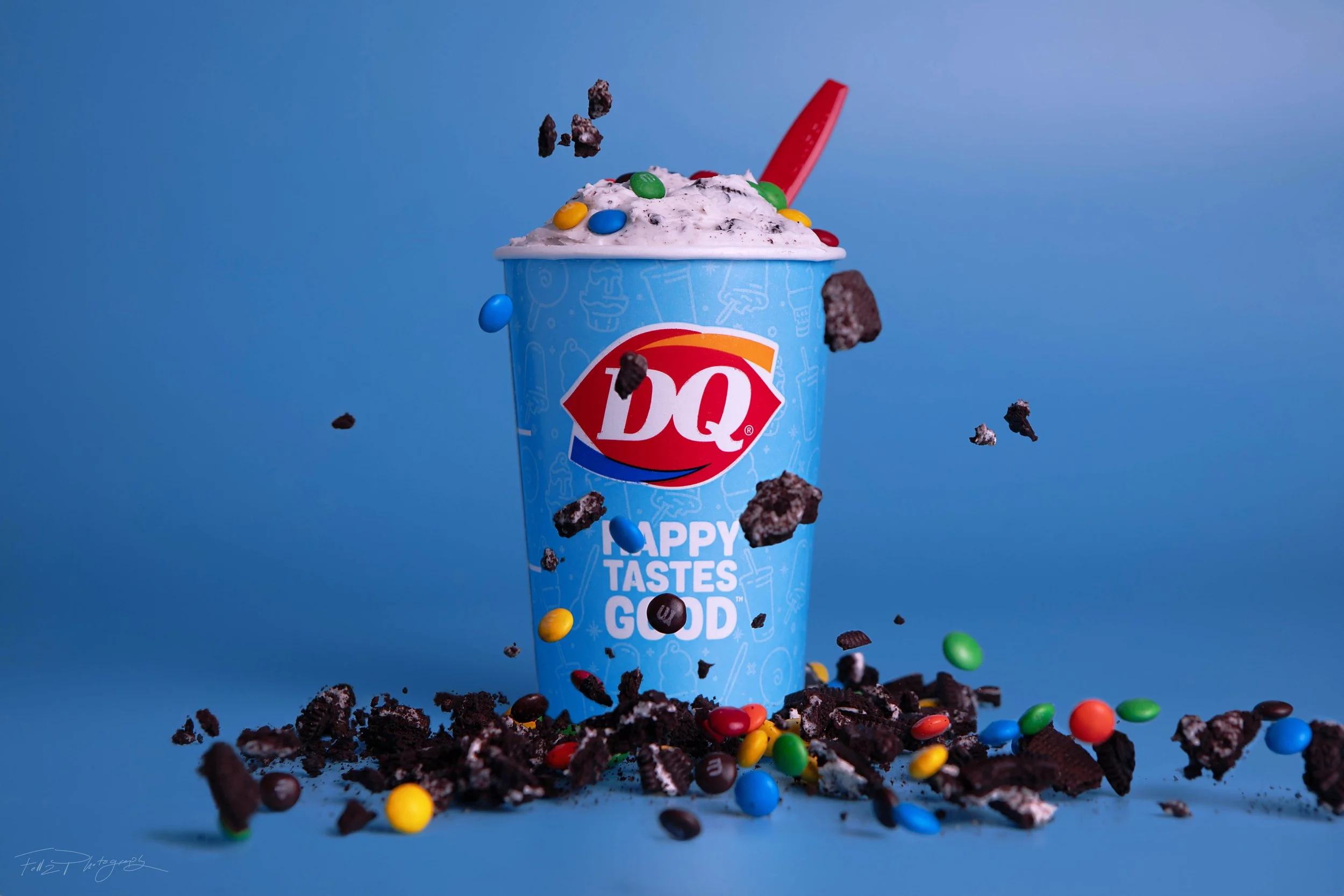 Dairy Queen ice cream in a blue cup with colorful candies and Oreo cookie pieces, with some toppings spilling out and in mid-air, against a blue background.