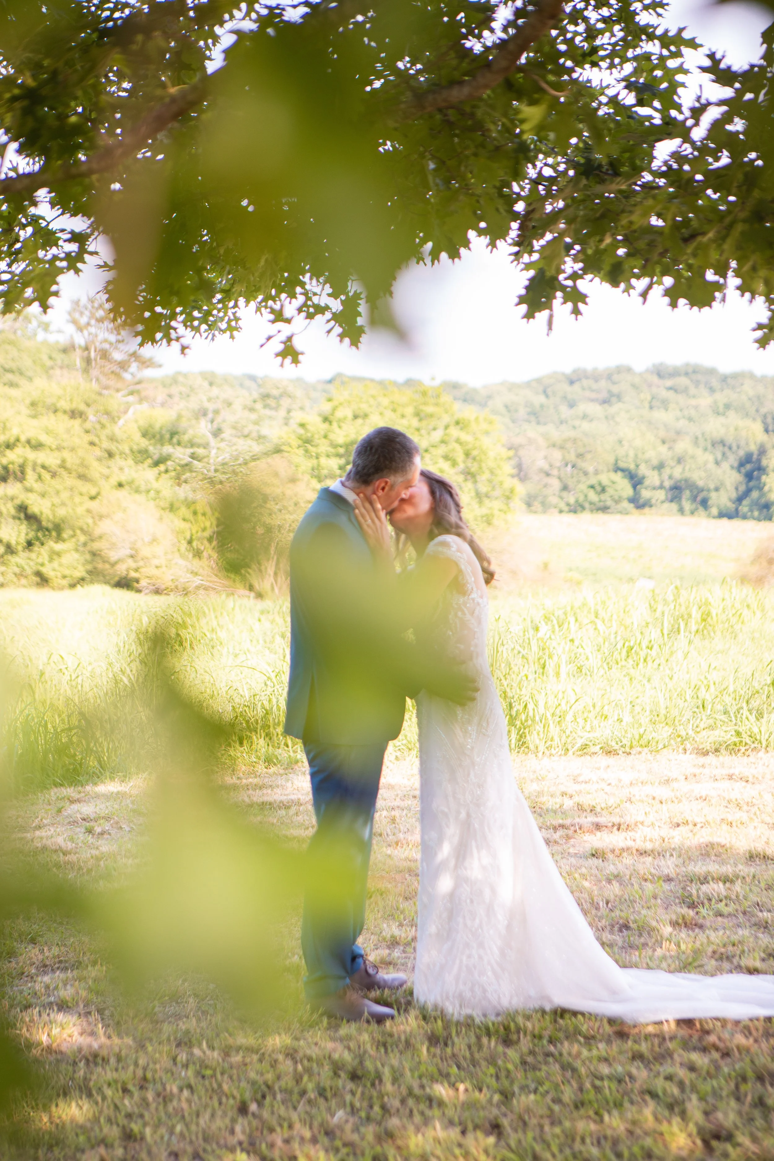 A couple sharing a kiss outdoors in a grassy field, with trees and hills in the background, framed by leaves in the foreground.