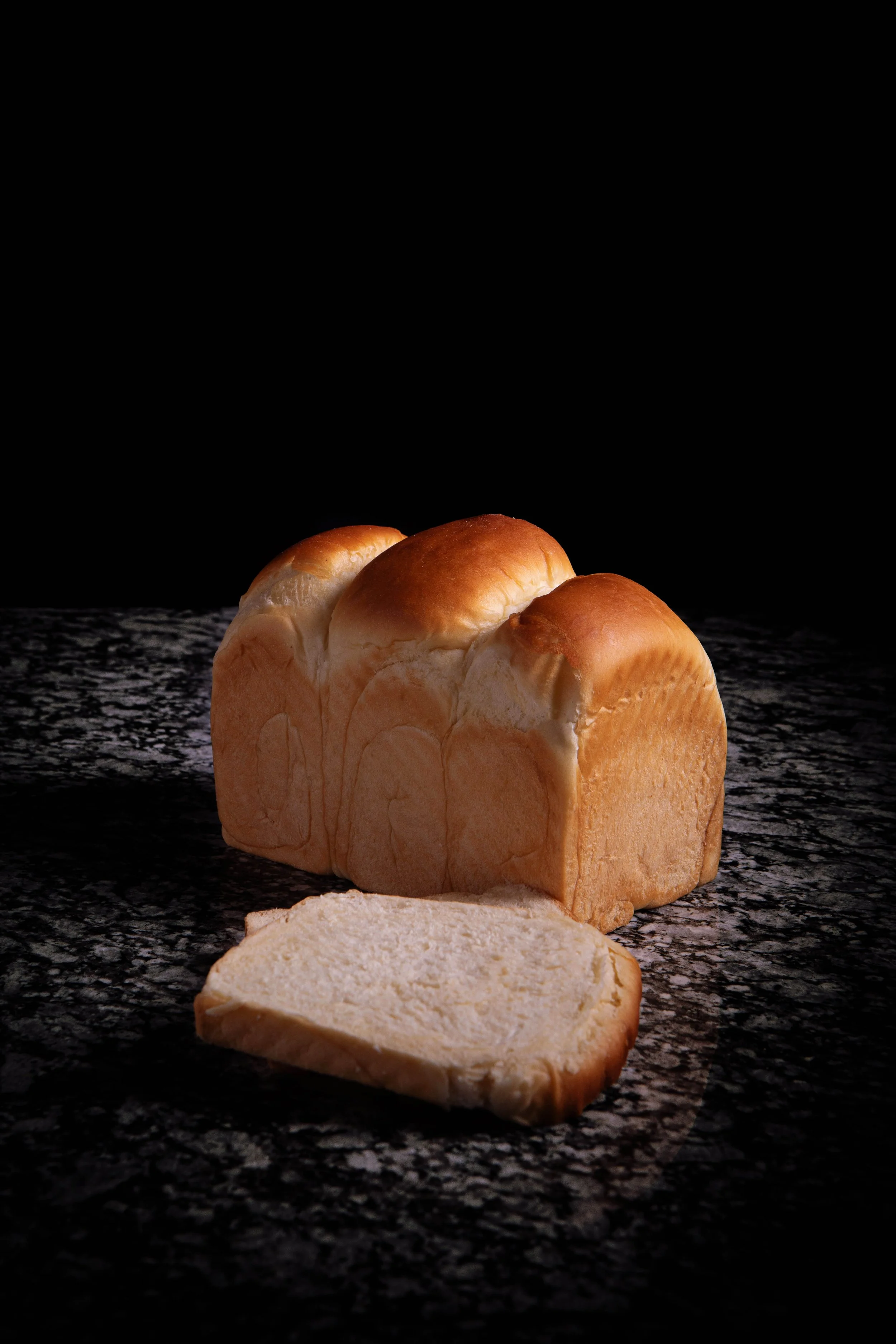Loaf of white bread with a slice cut, on a dark speckled surface against a black background